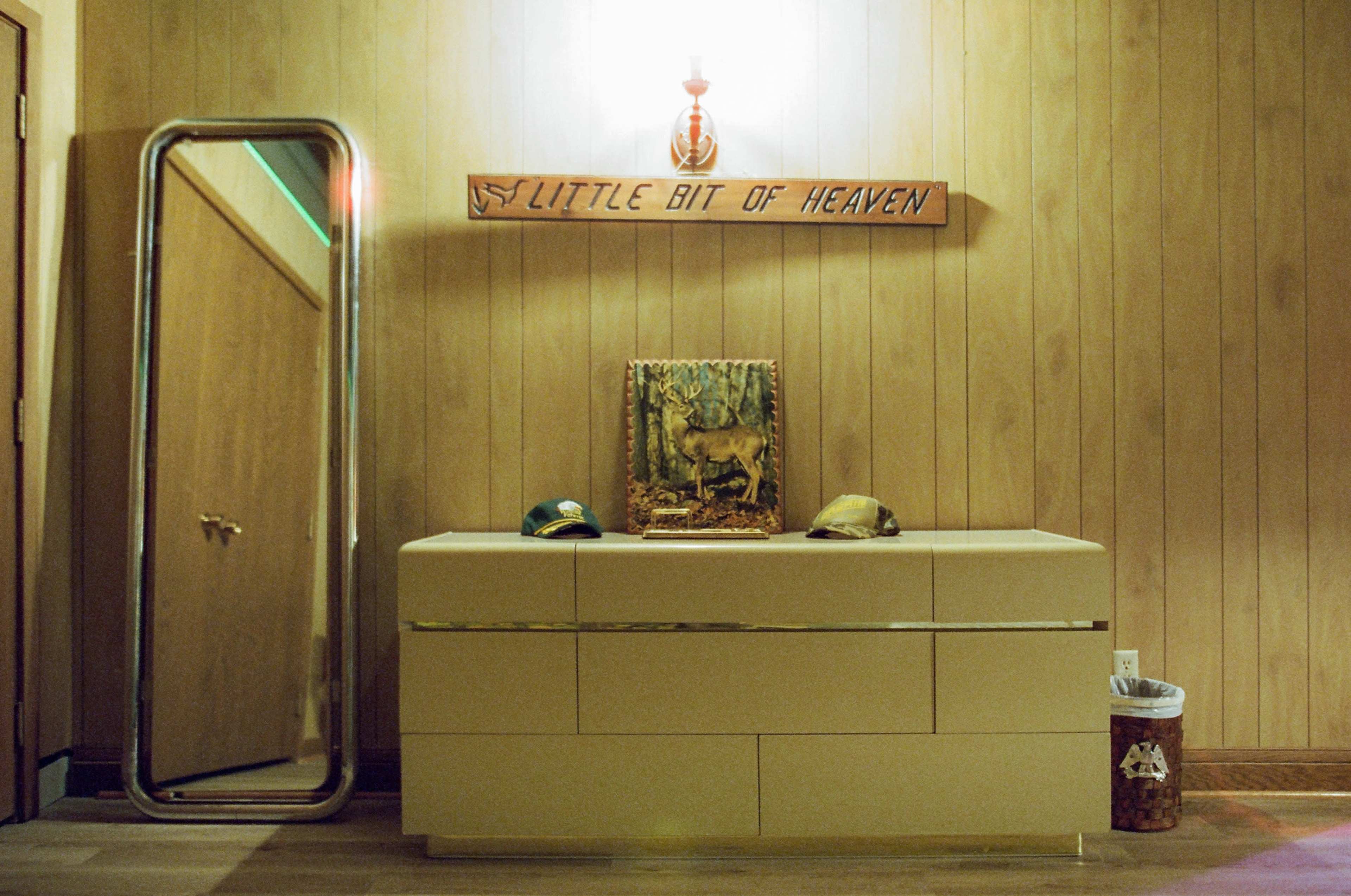 A wooden dresser with two hats and a framed picture of a deer is positioned beneath a wall-mounted light in a room with paneled wood walls.