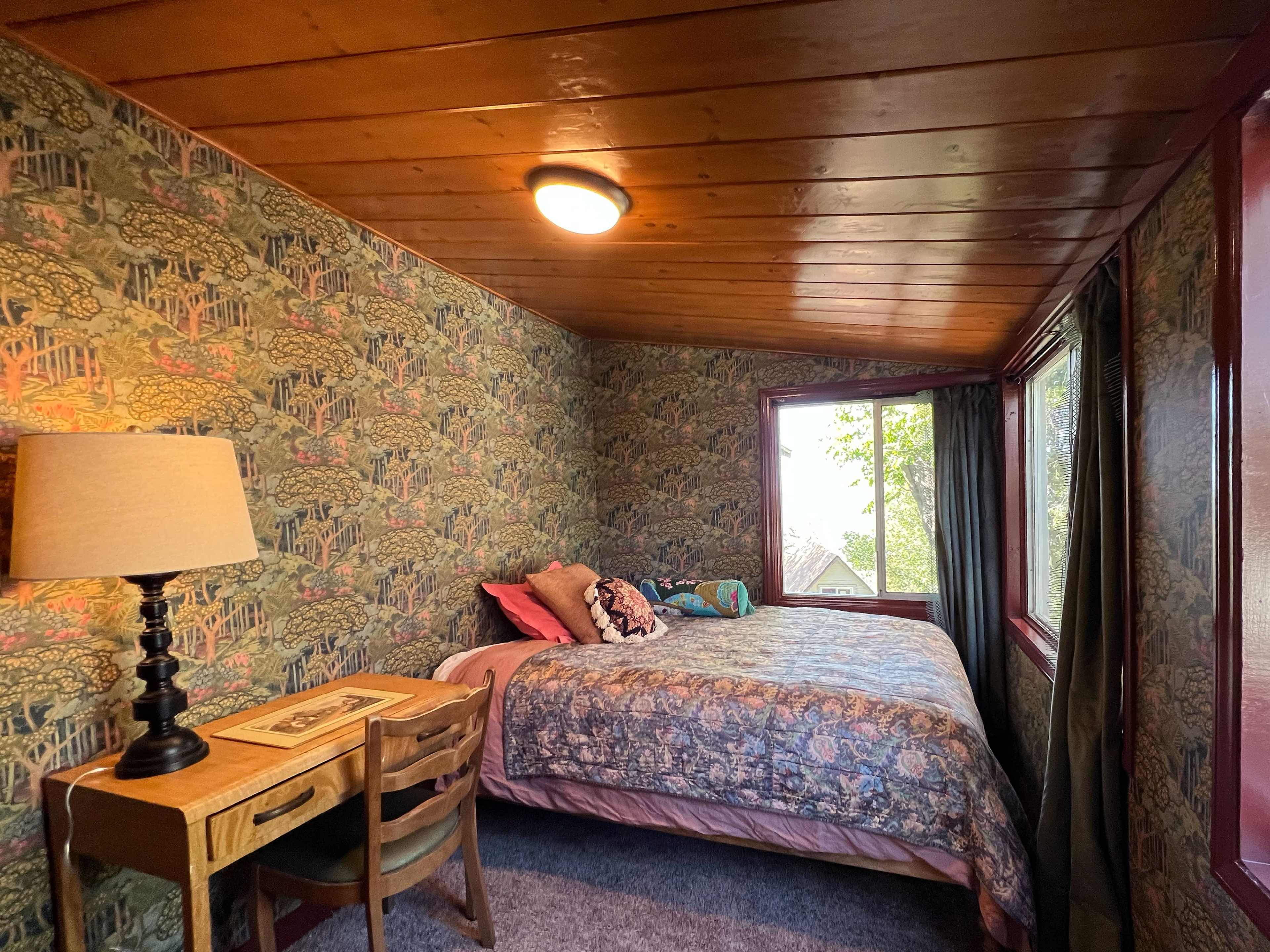 A bedroom with patterned wallpaper, a bed with decorative pillows, a small table, and windows overlooking greenery.