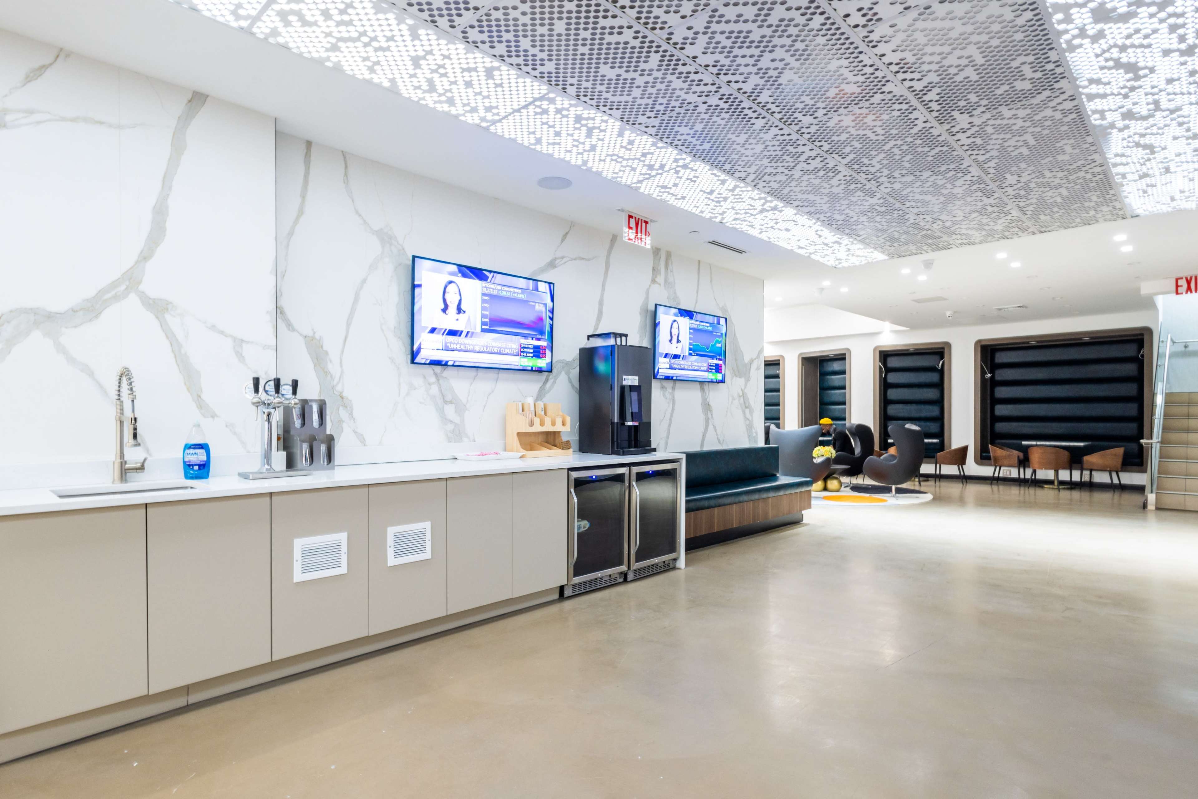 The image shows a modern office lounge area with a kitchenette, two televisions mounted on the wall, and a seating area featuring a dark couch and colorful chairs.