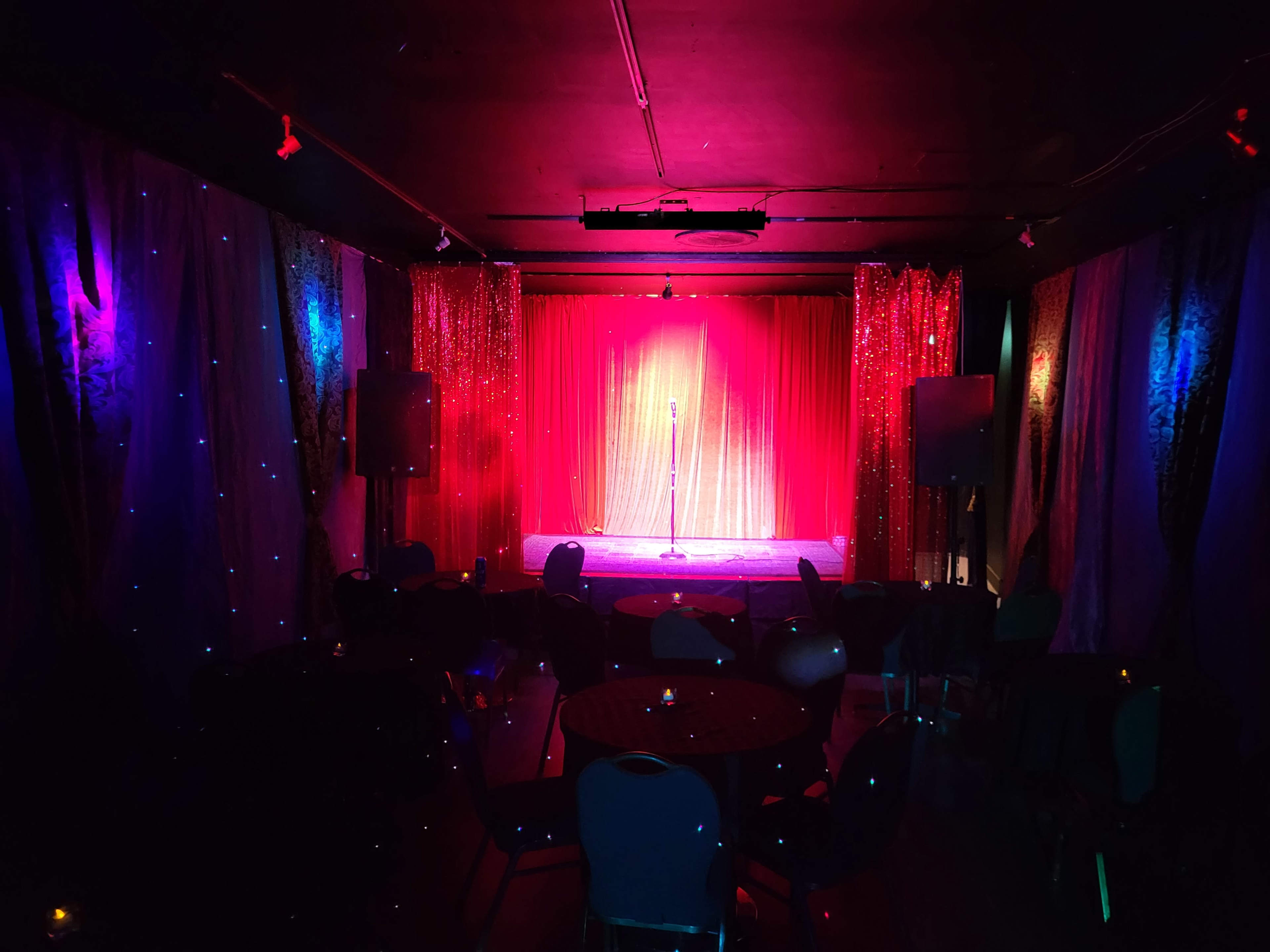A dimly lit performance stage with a microphone at center, surrounded by colorful fabric drapes and empty tables arranged for an audience.
