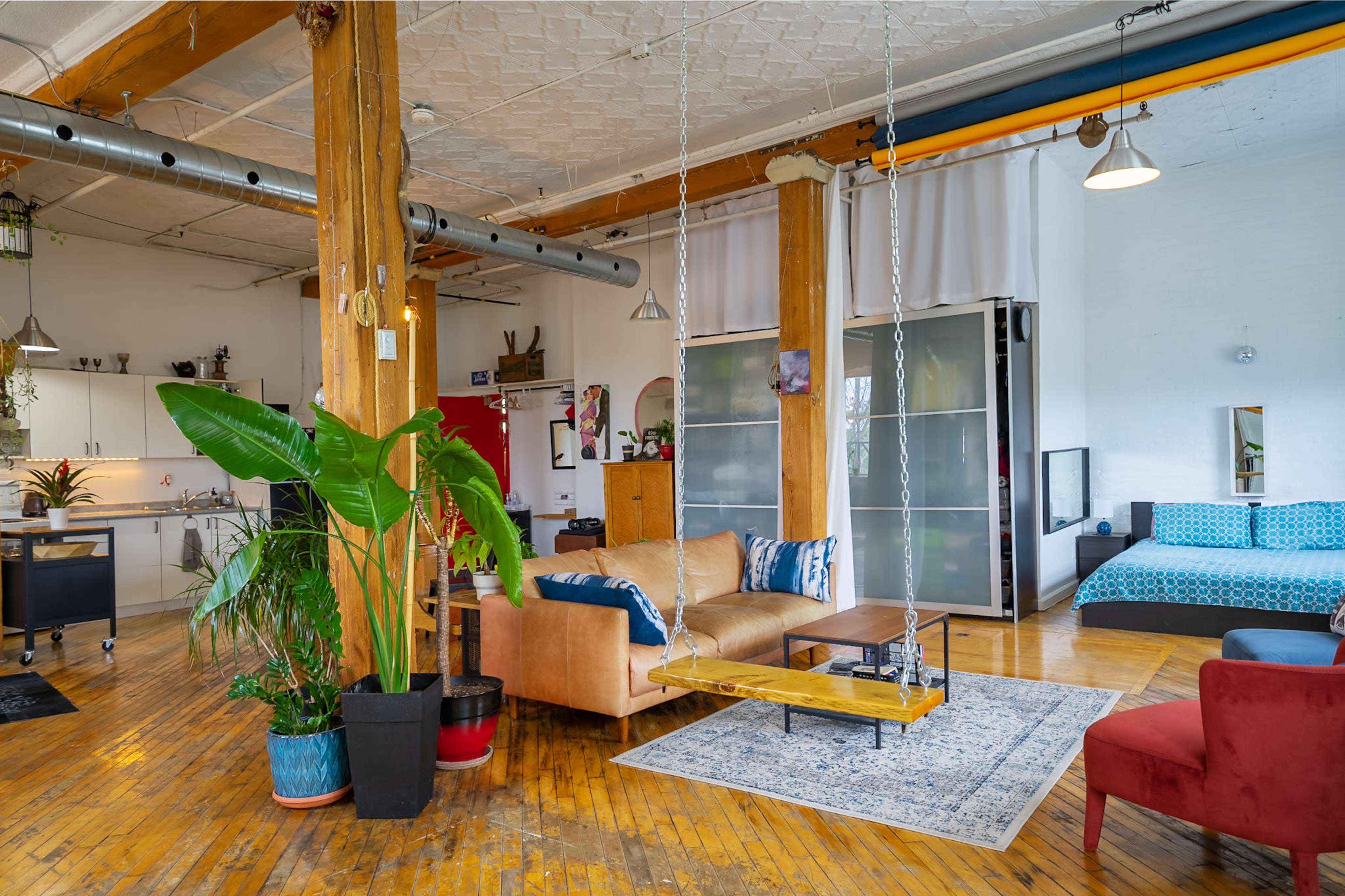 The image shows a spacious, open living area with wooden floors, featuring a mix of seating options, a large plant, and sliding glass doors.