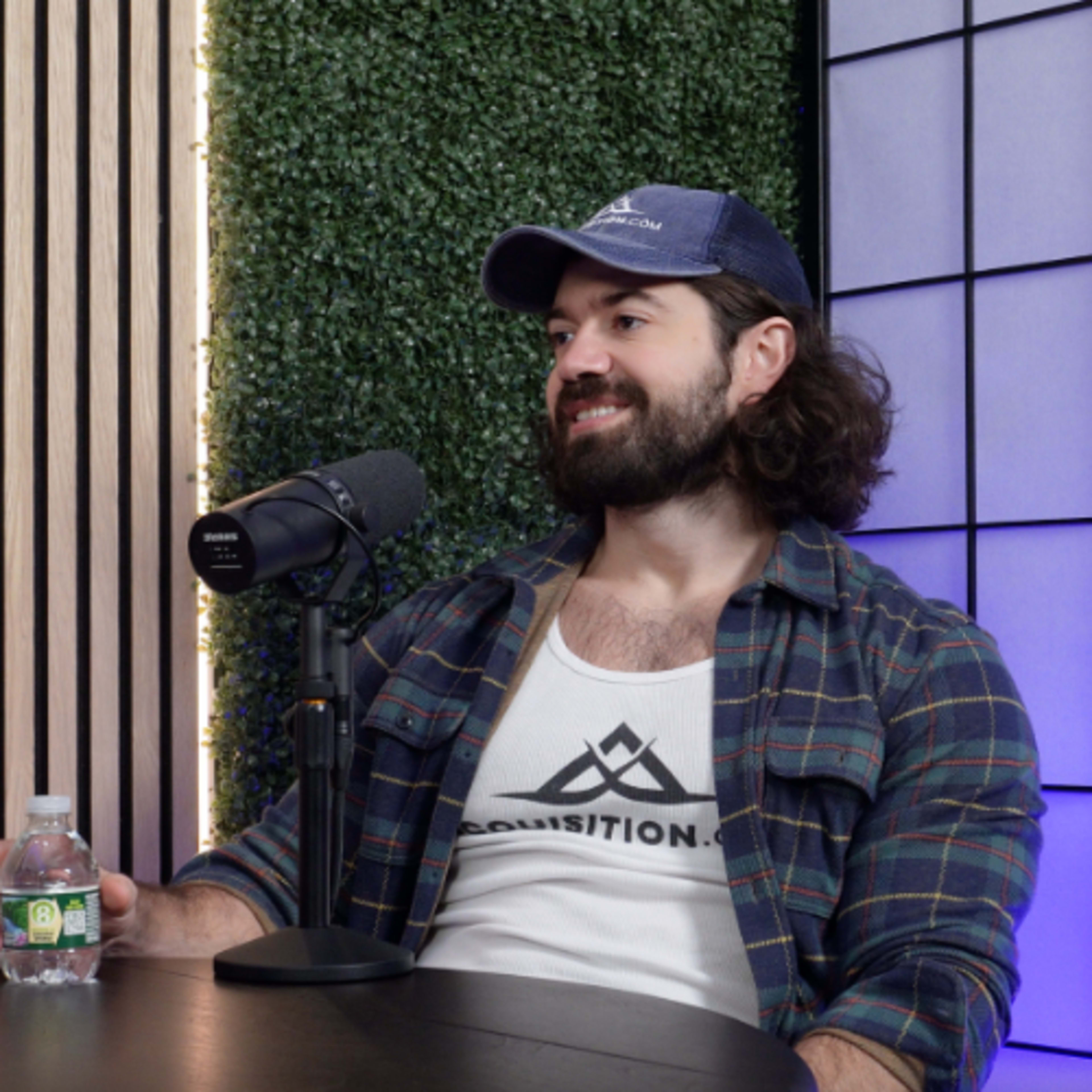 A man wearing a cap and a tank top sits at a table with a microphone and a water bottle, in front of a backdrop of greenery and lights.