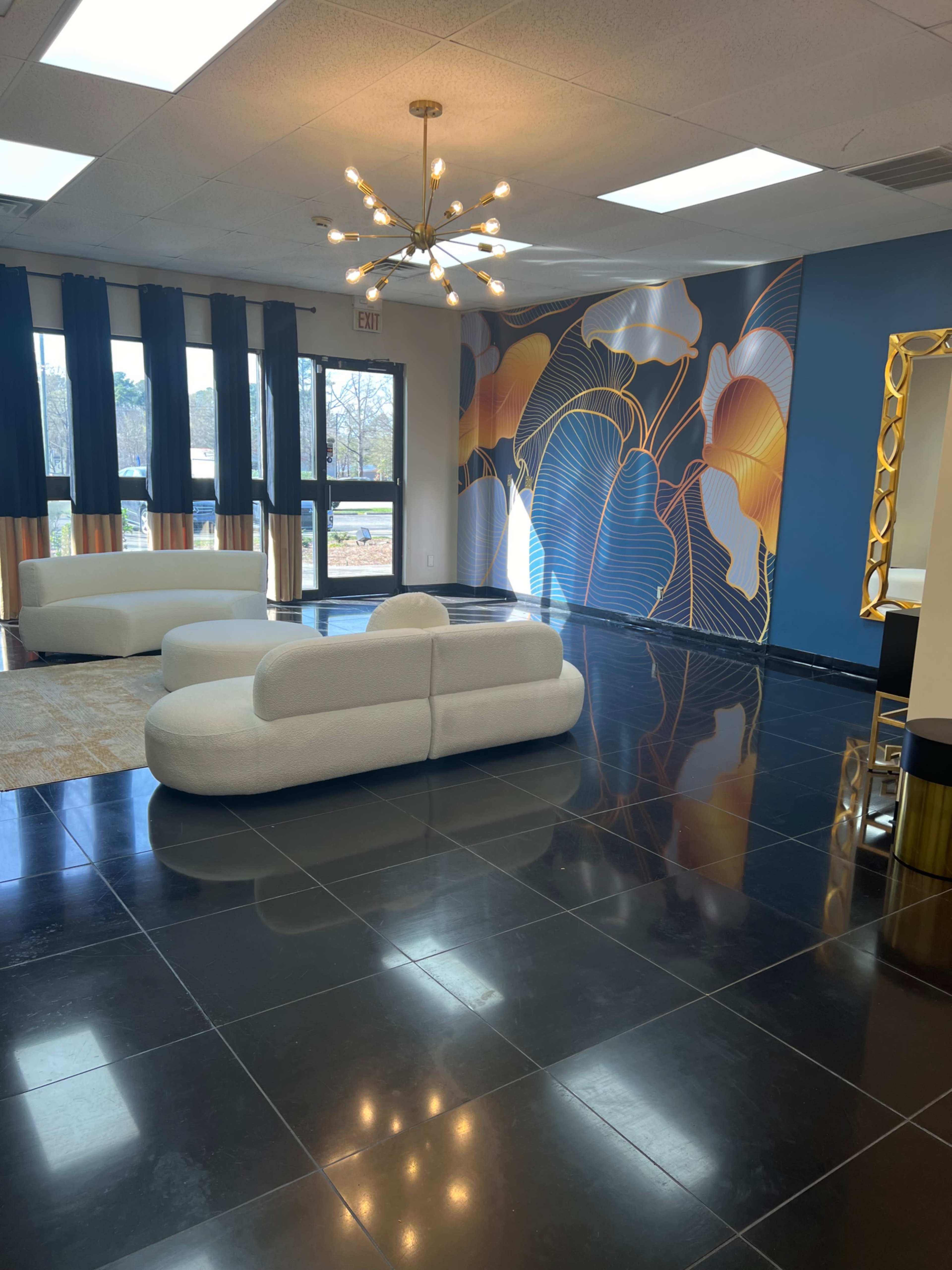 A spacious room with black tiled flooring, featuring a white sofa, a large colorful mural on the wall, and ample natural light coming through large windows.