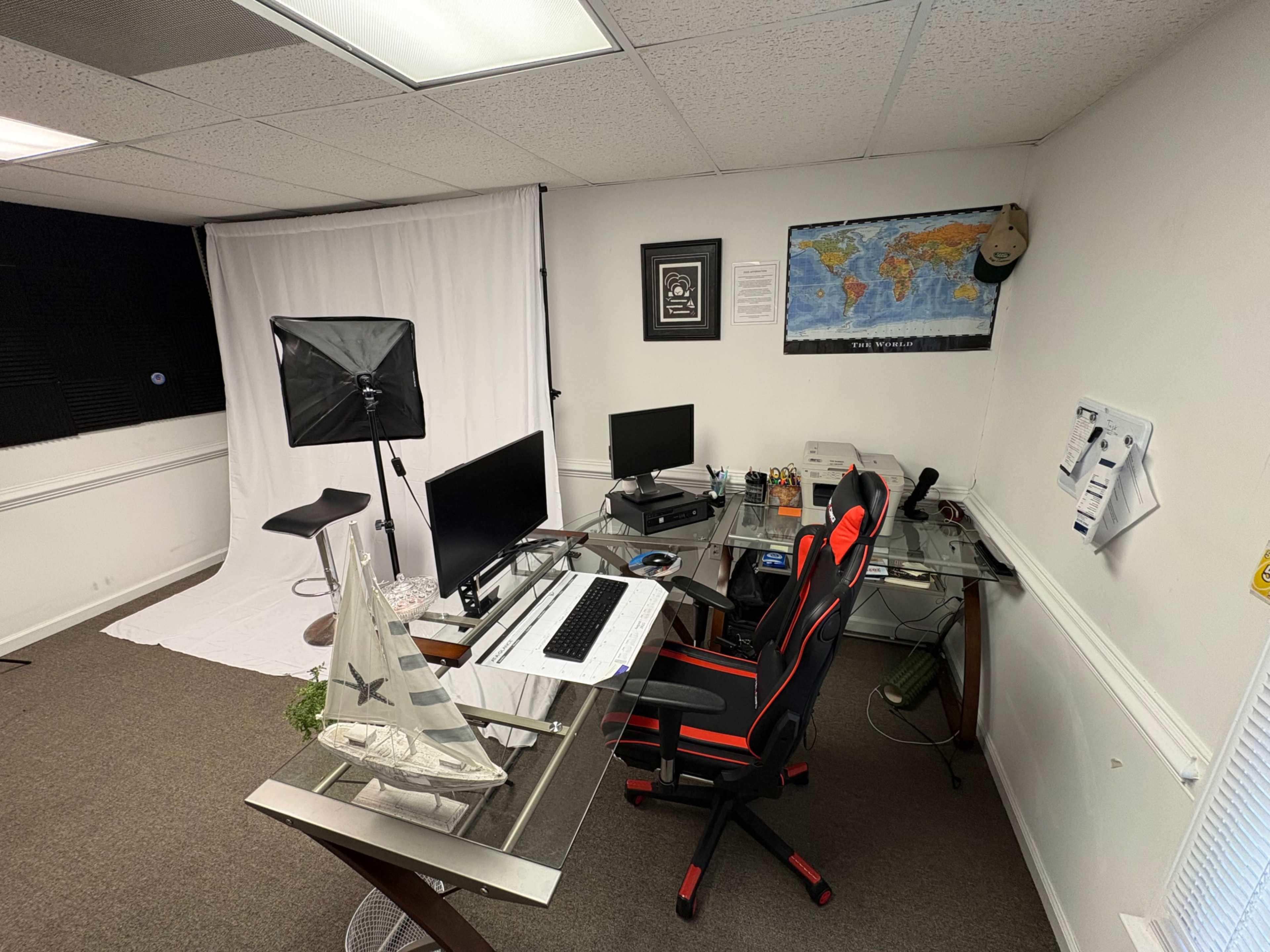 The image shows a modern office space with two computer monitors, a desk equipped with a keyboard, a camera setup featuring a backdrop, and a map on the wall.