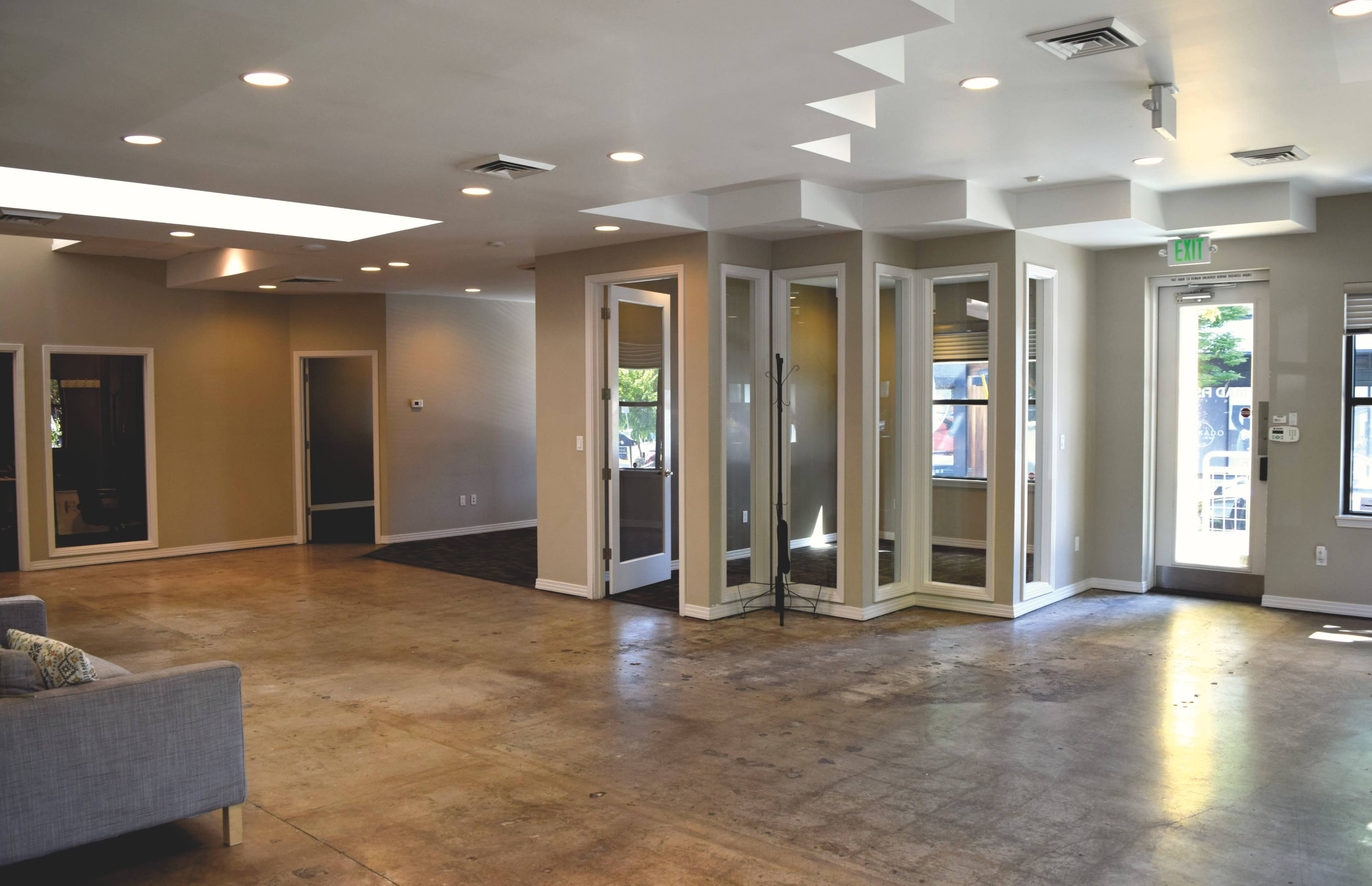 The image shows a spacious interior of a modern commercial space featuring concrete flooring, large windows, and several glass-enclosed rooms.
