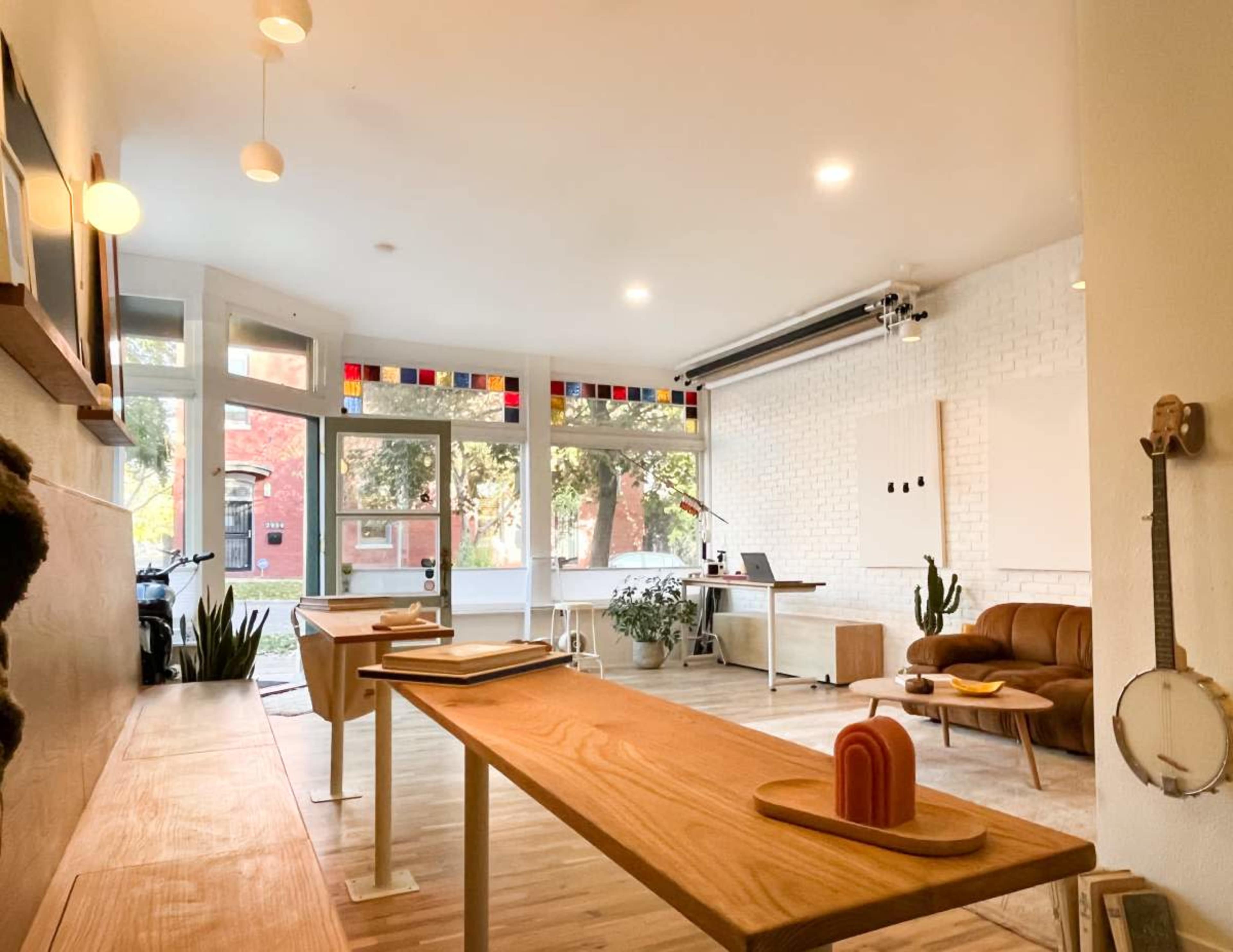 A modern, well-lit workspace with wooden tables, a brown couch, and large windows featuring colorful stained glass.