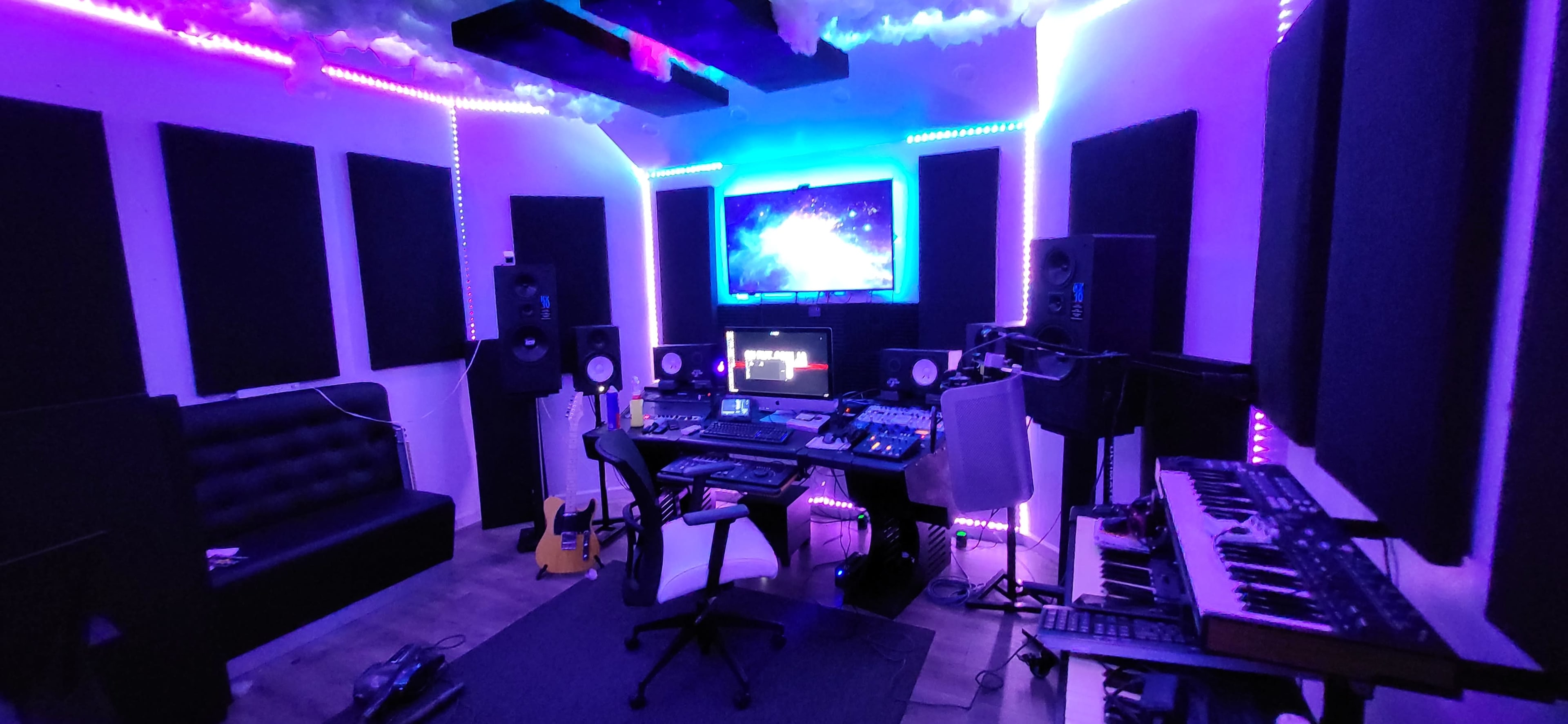 A music production studio features a central workstation with multiple screens, audio equipment, and various instruments, surrounded by sound-absorbing panels and LED lighting.