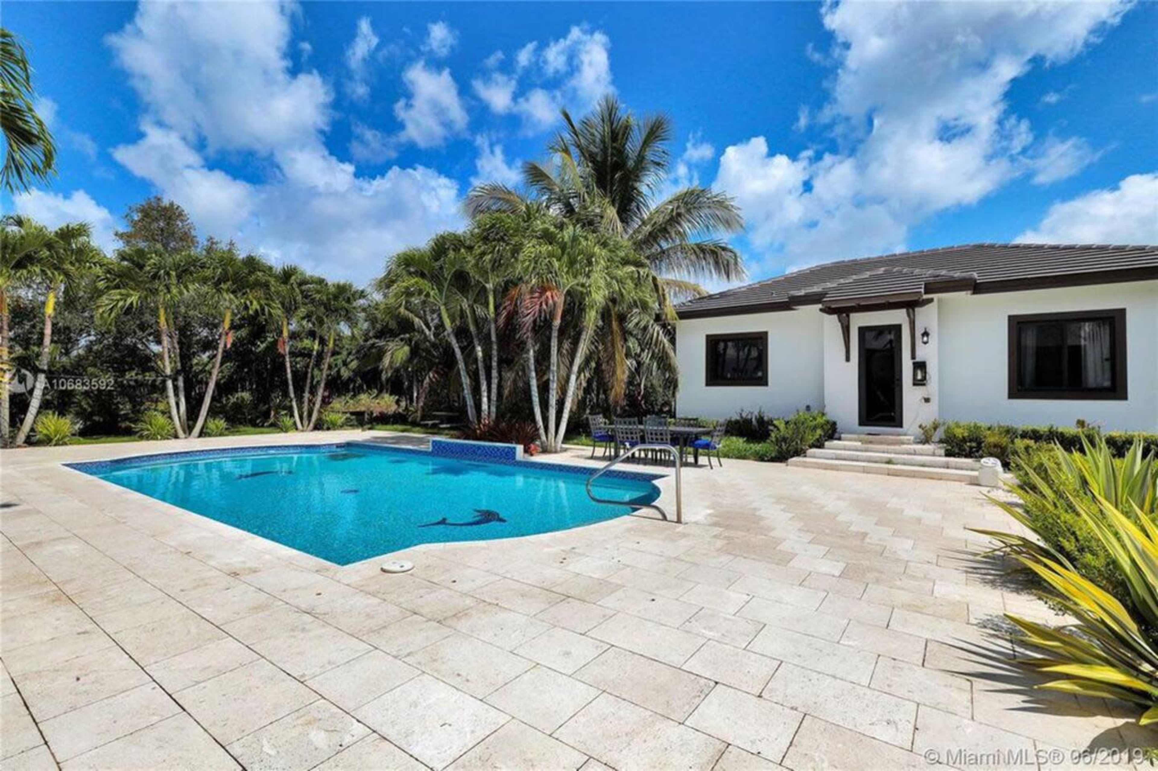 A residential backyard with a swimming pool surrounded by palm trees and a house in the background.