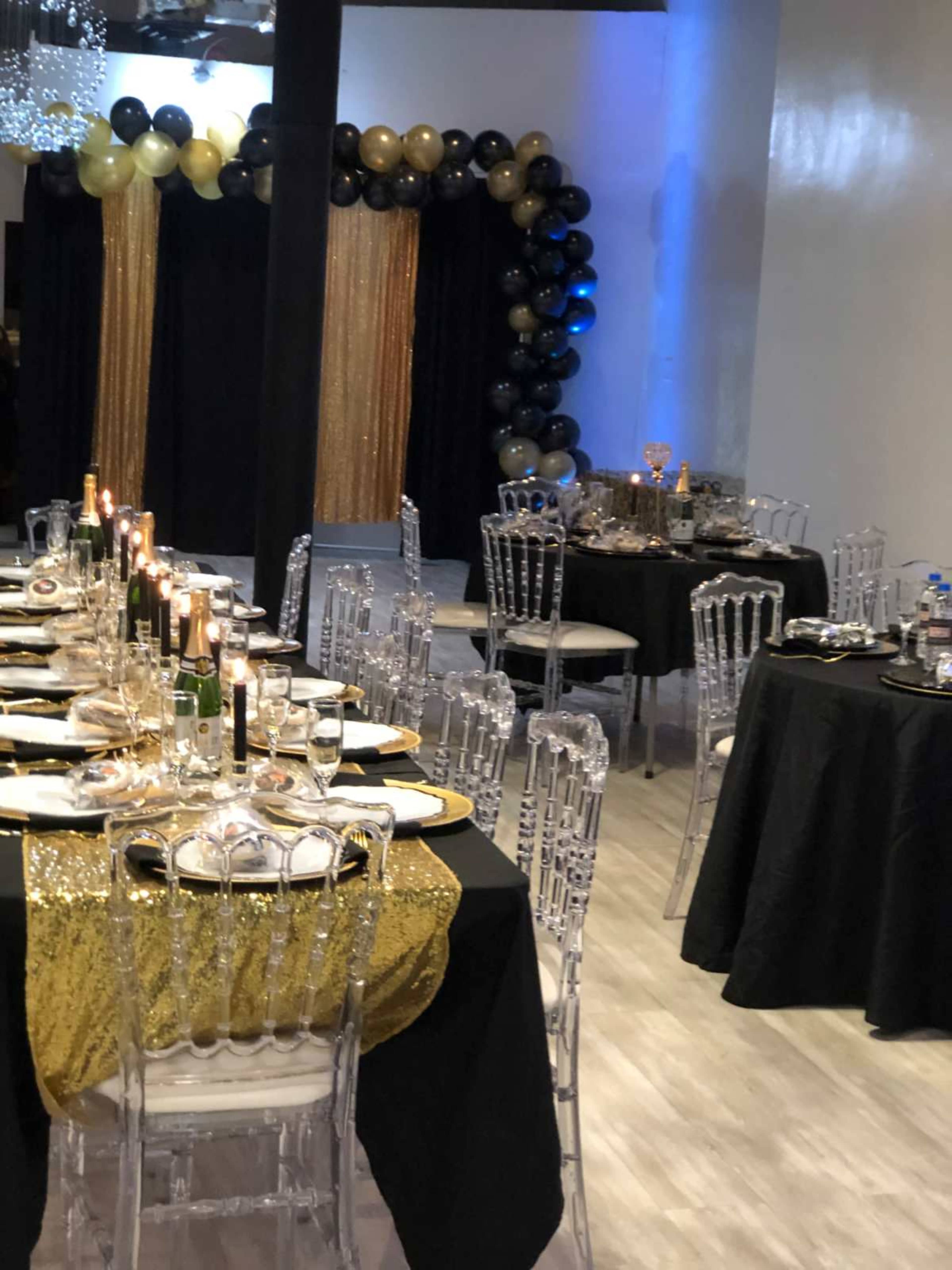 The image shows a decorated event space with elegant table settings, featuring clear chairs, black and gold tablecloths, and a backdrop of balloons.
