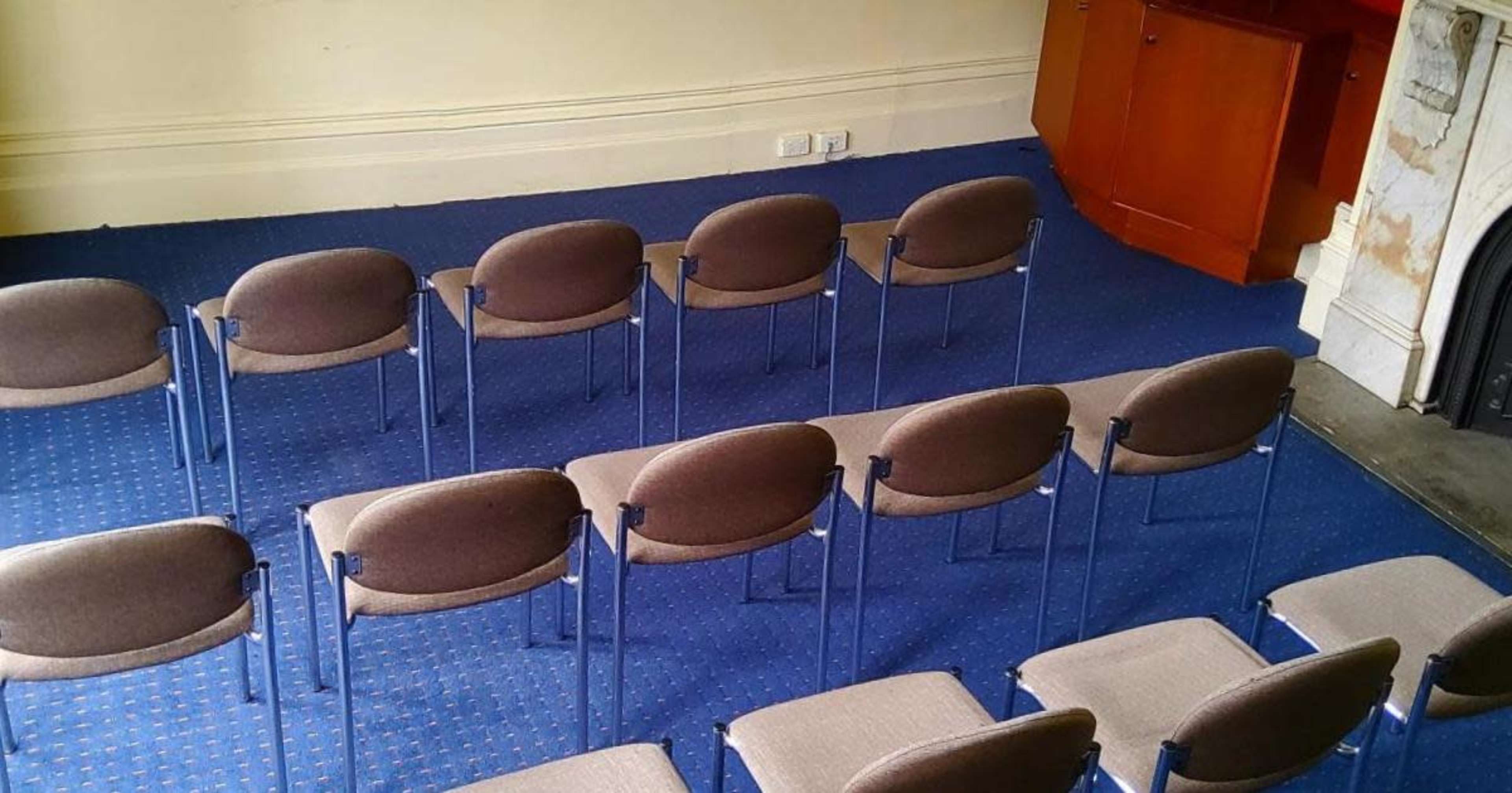 A set of brown chairs is arranged in rows on a blue carpet in a room with a wooden cabinet and a fireplace.