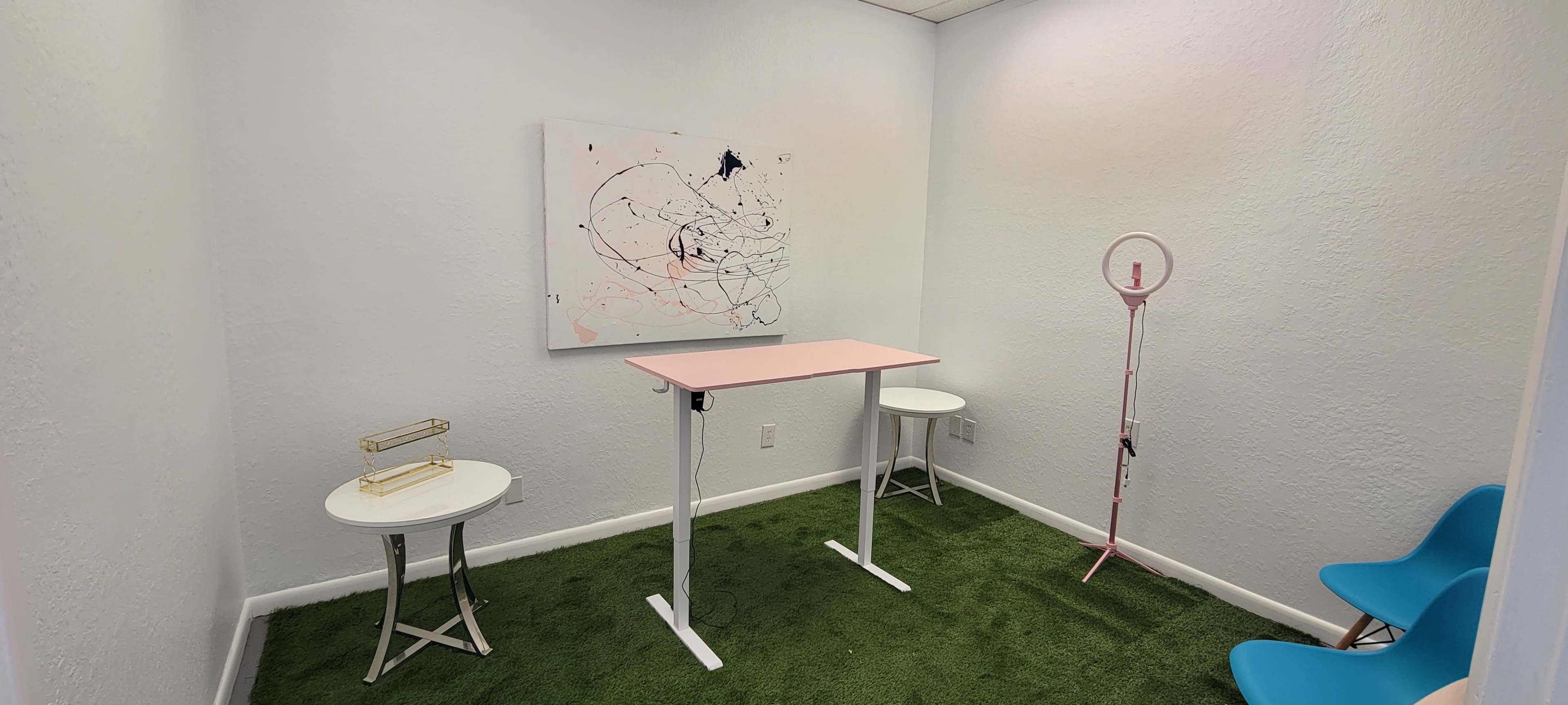 The image shows a small, minimalist room with light blue walls, green carpet, a pink standing desk, a small white table, and a ring light on a stand.
