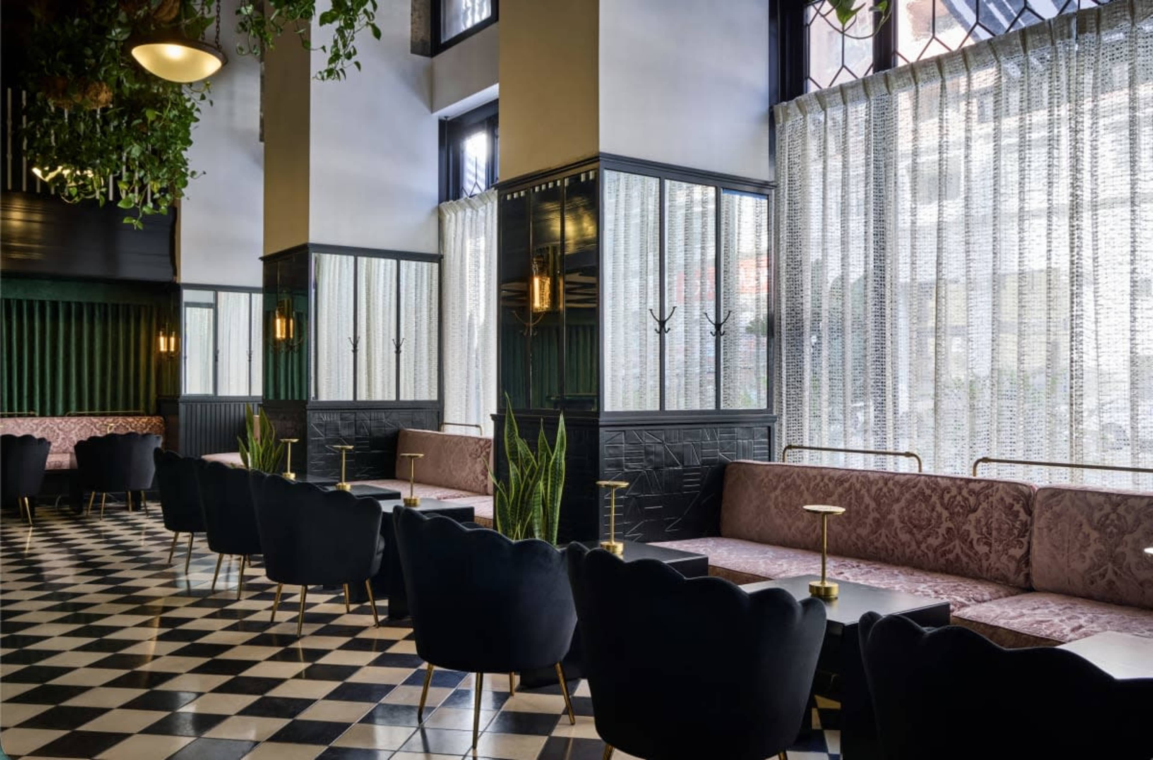 The image depicts a modern café with stylish seating arrangements, large windows covered by sheer curtains, and a checkered floor pattern.
