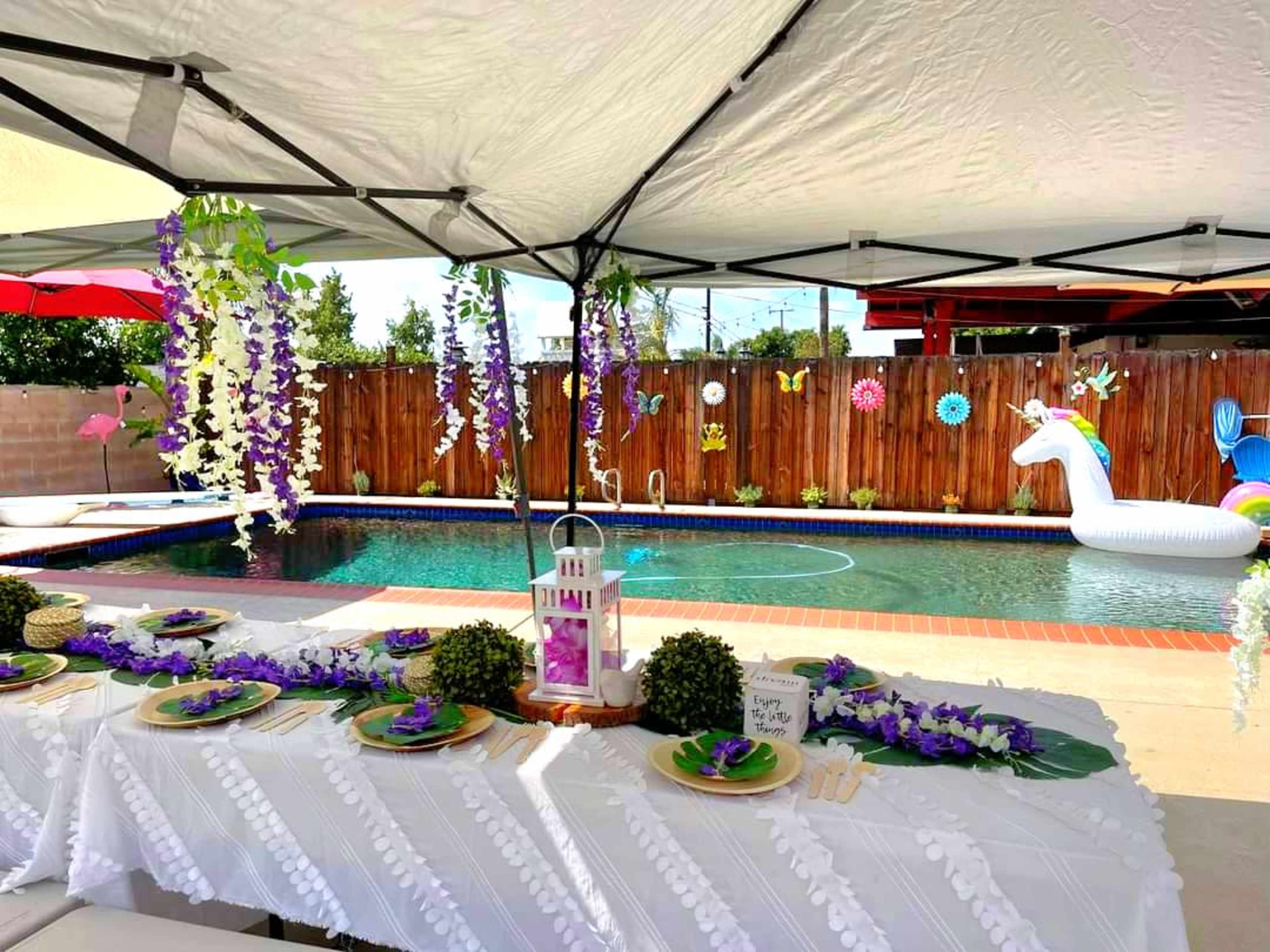 A decorated outdoor space features a table set with floral arrangements and a swimming pool in the background, complete with a unicorn float and hanging decorations.