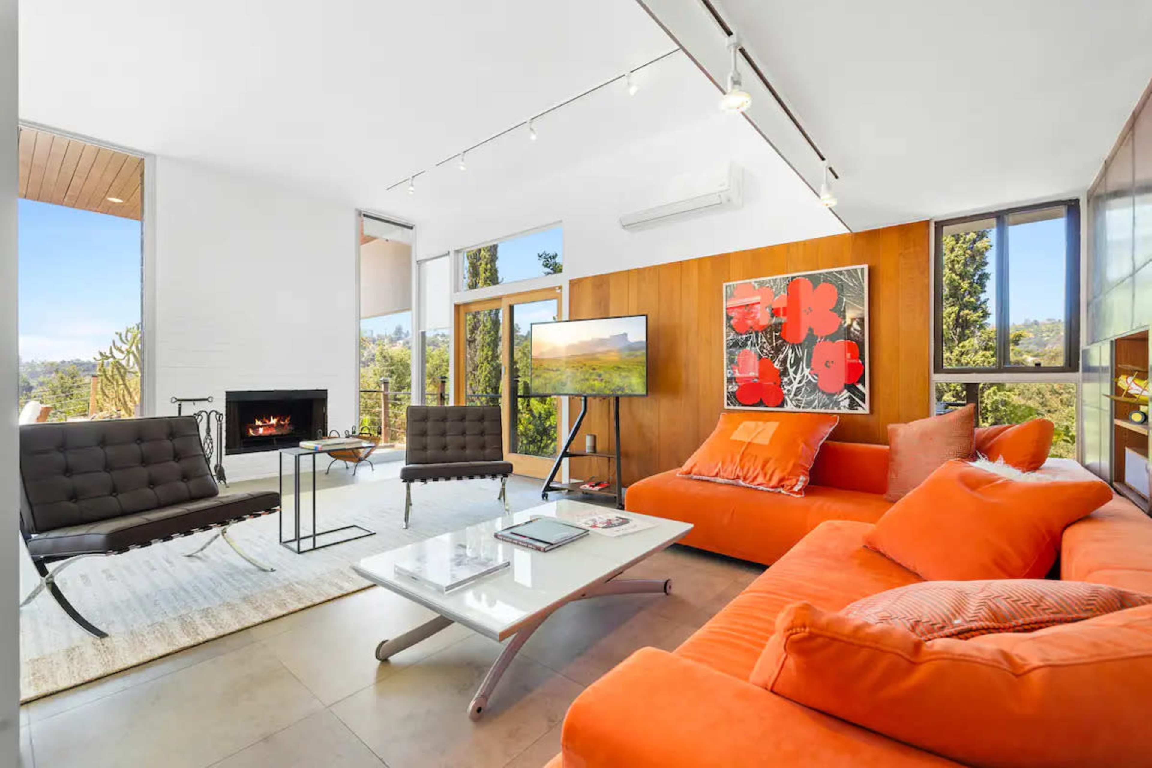 The living room features an orange sectional sofa, black chairs, a glass coffee table, and a television, with large windows overlooking a scenic view.