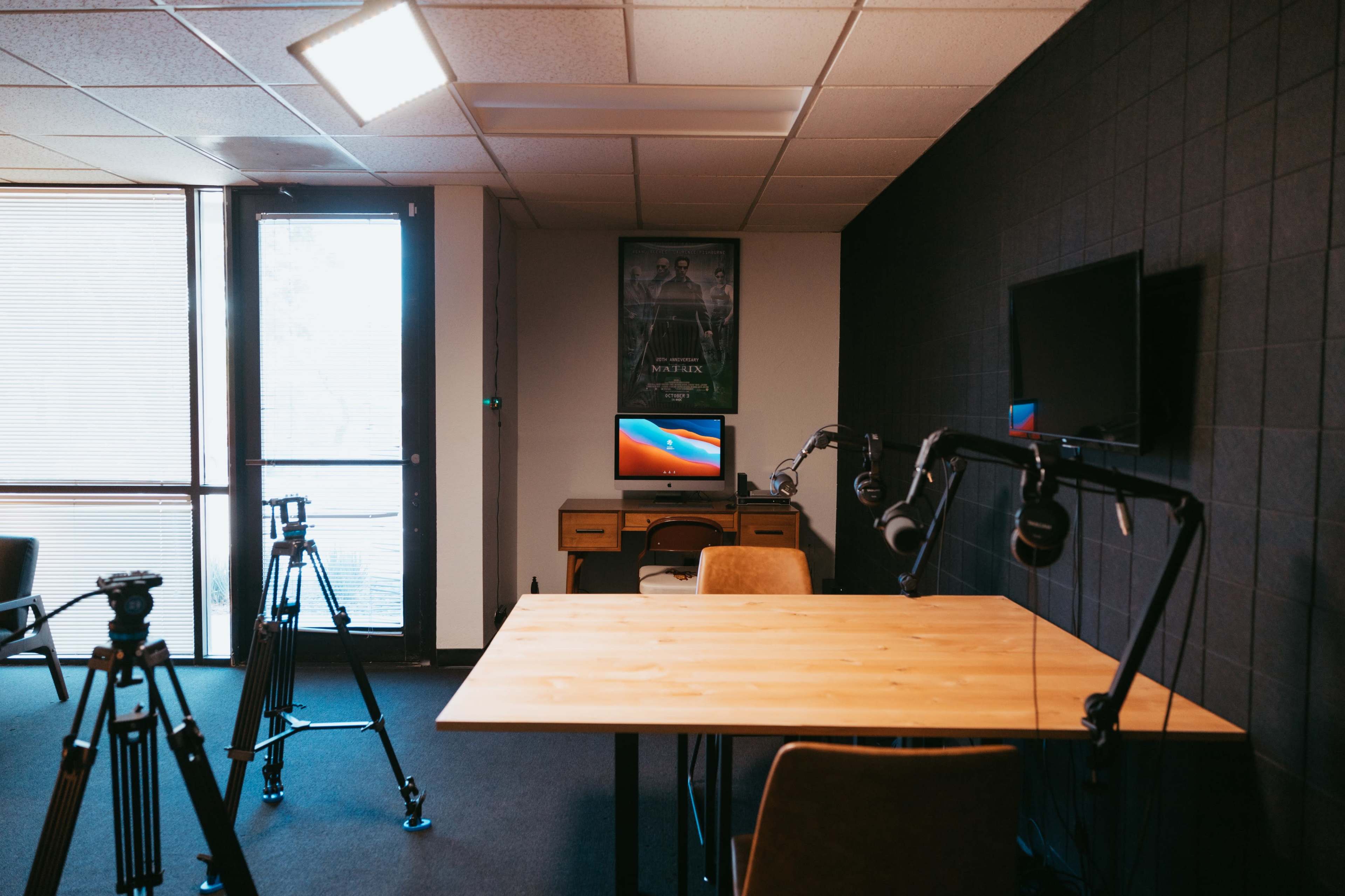 A modern recording studio with a wooden table, chairs, and camera tripods set up near a large window and a wall-mounted TV.