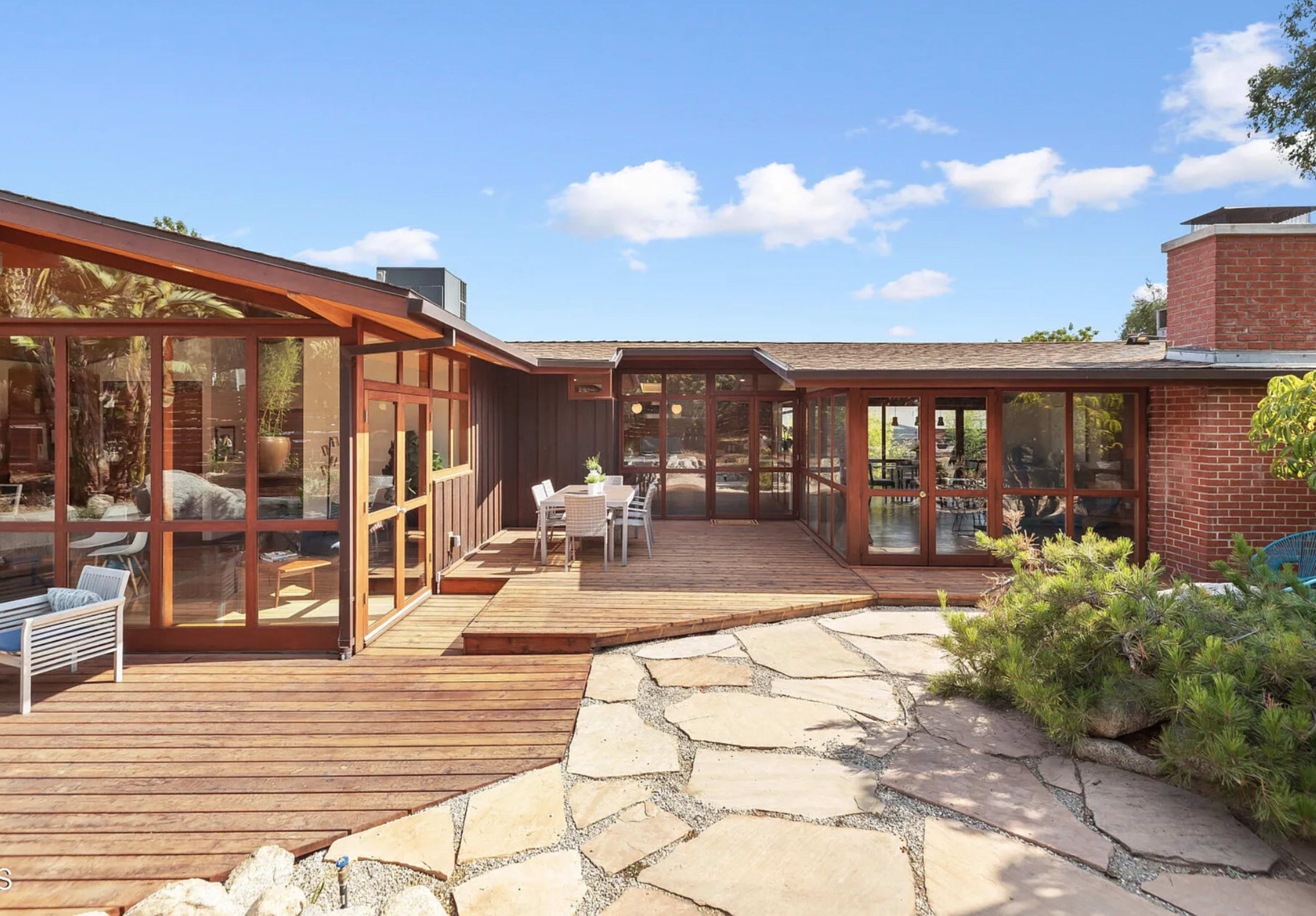 The image shows a modern single-story home with large glass windows and a stone patio surrounded by greenery.
