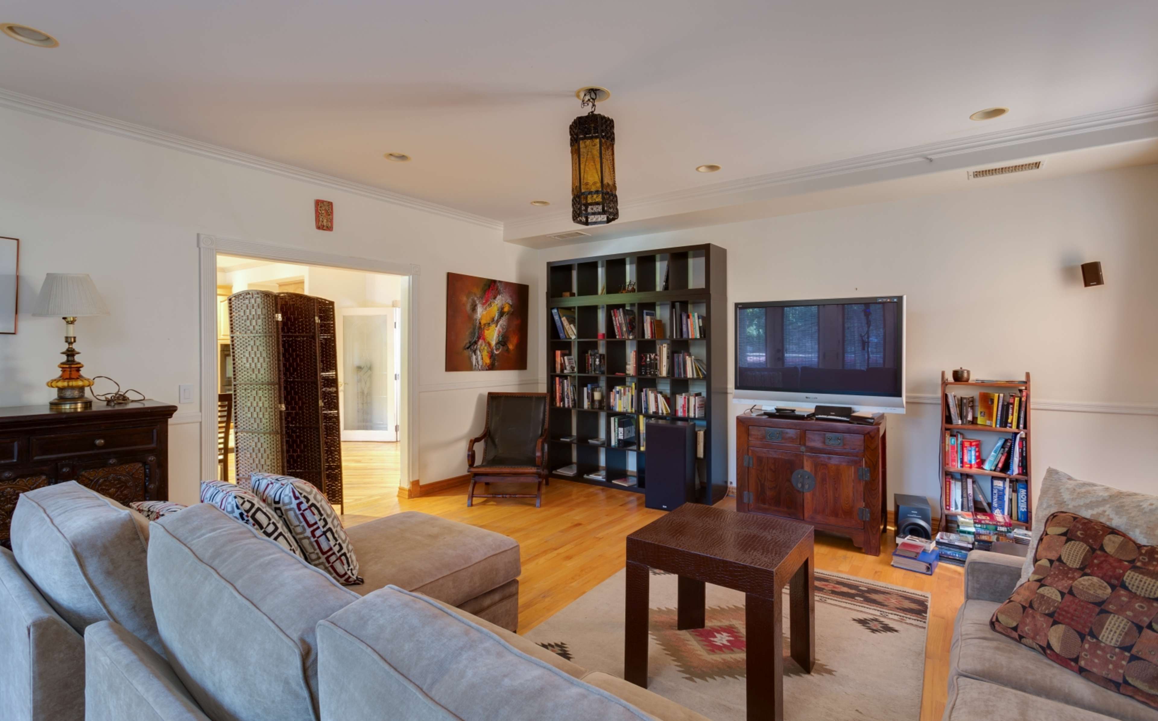 A cozy living room features a large sectional sofa, a wooden coffee table, a bookshelf filled with books, and a flat-screen TV, with wood flooring and ambient lighting.