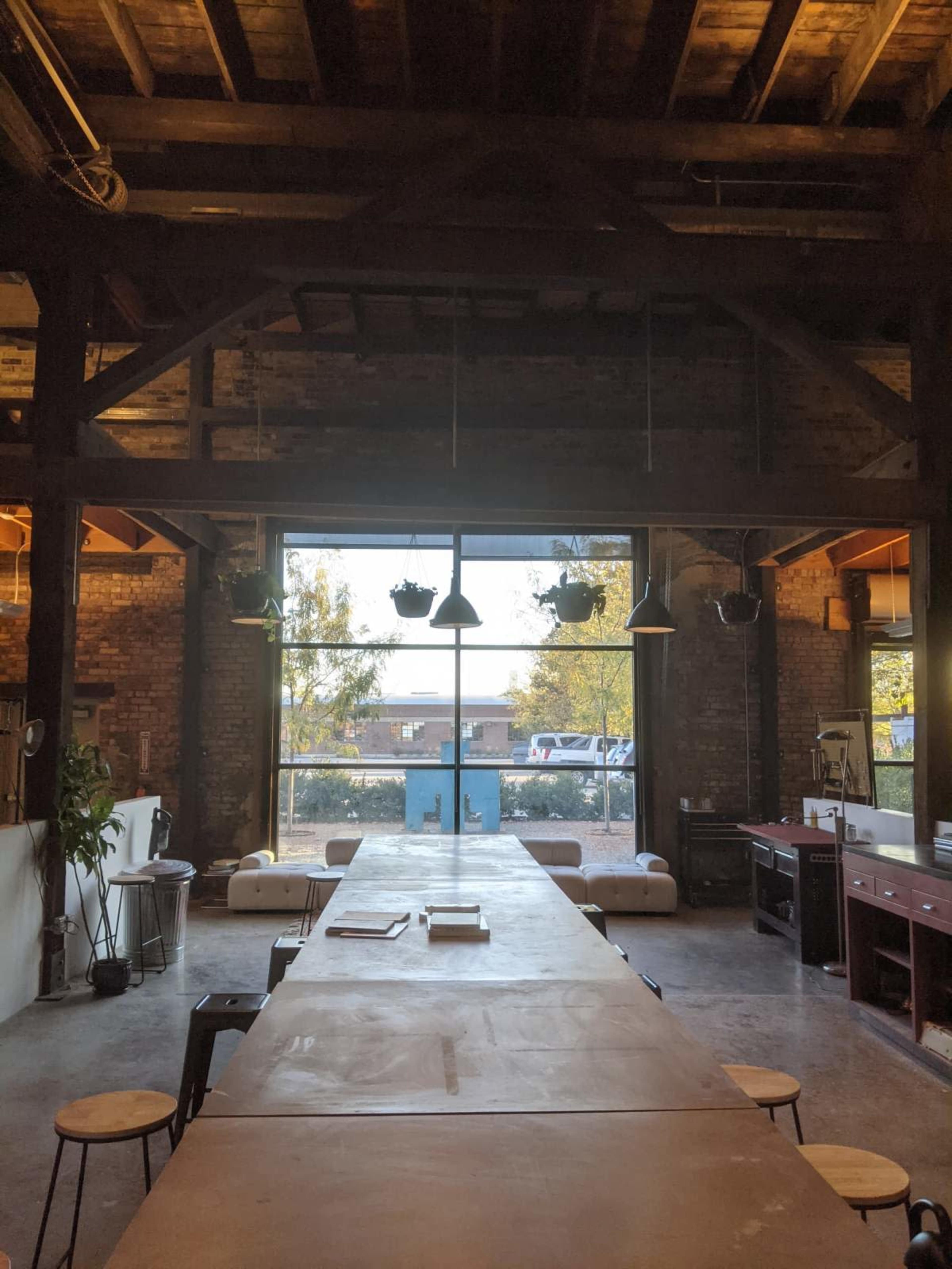 A long wooden table with stools is positioned in front of large windows overlooking an industrial space with exposed brick walls and ceiling beams.
