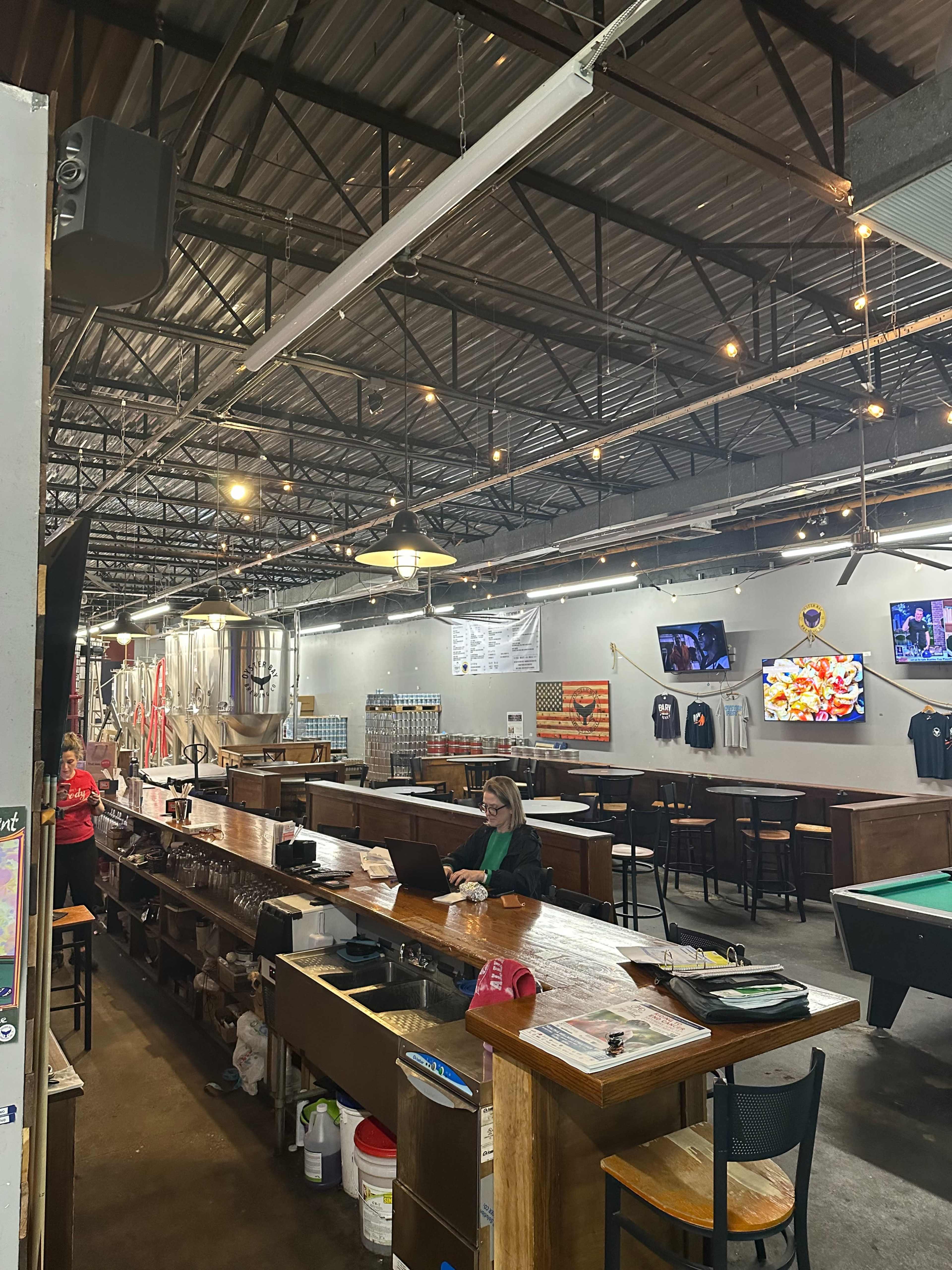 The image shows the interior of a bar with a long wooden counter, tables, and bar stools, featuring multiple televisions mounted on the walls.