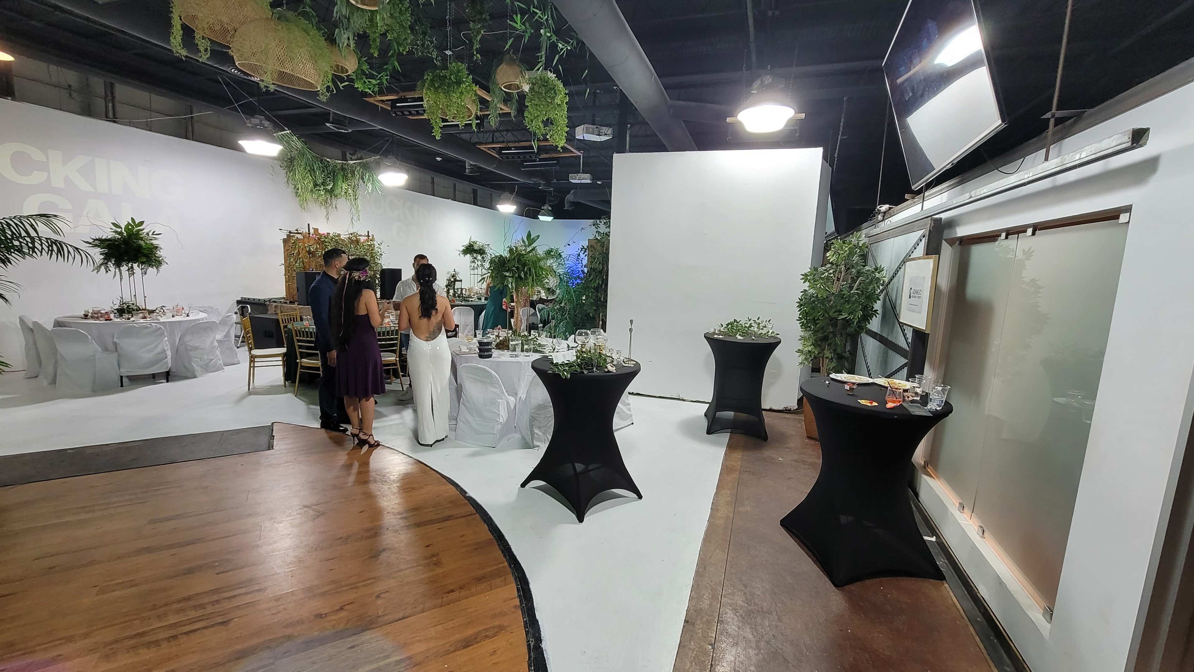 A spacious event venue with tables set for a gathering, featuring white and black decor elements and greenery accents.