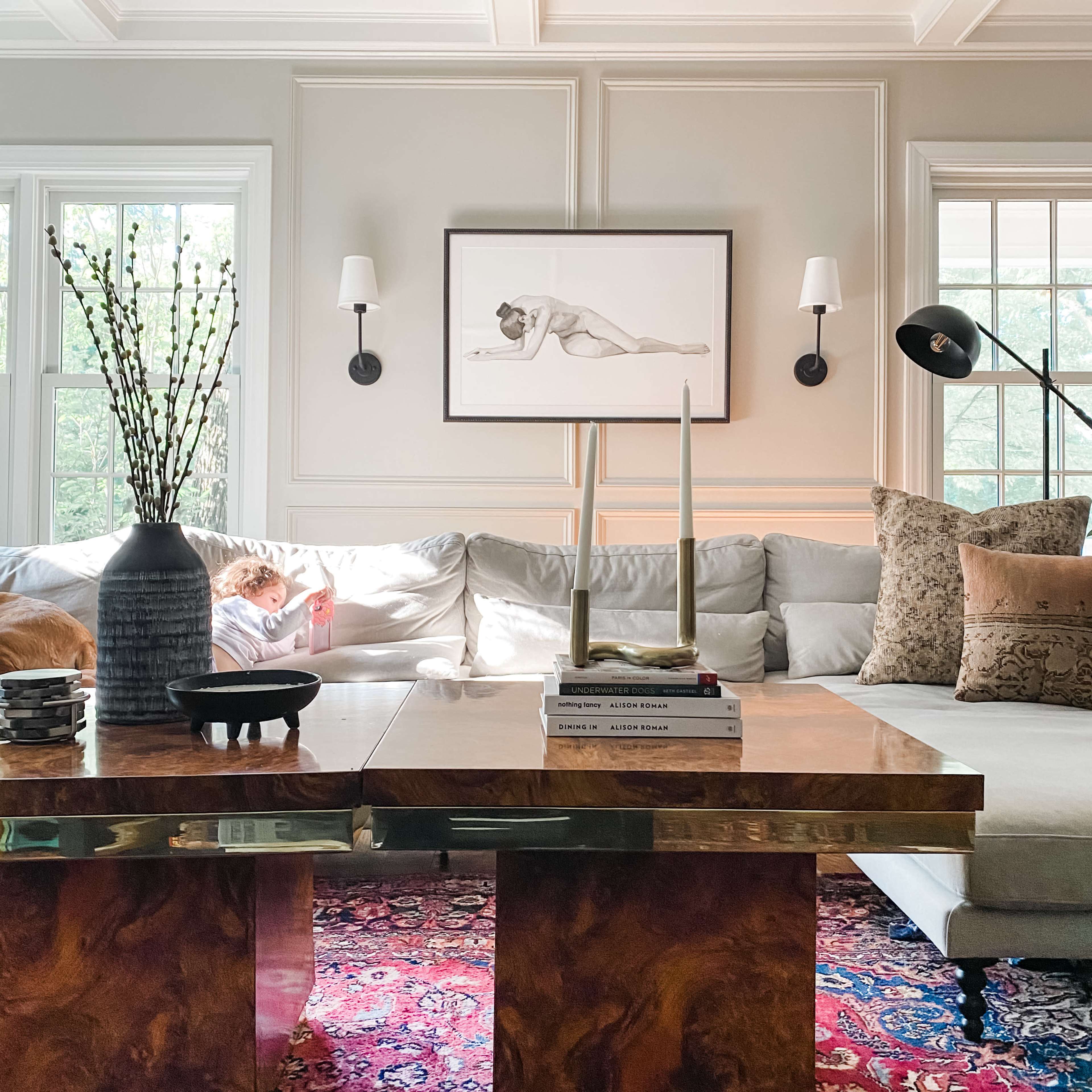 A bright living room features a large sofa with decorative pillows, a coffee table with books and a decorative bowl, and a piece of artwork on the wall, while a person is seated in the background reading on a mobile device.
