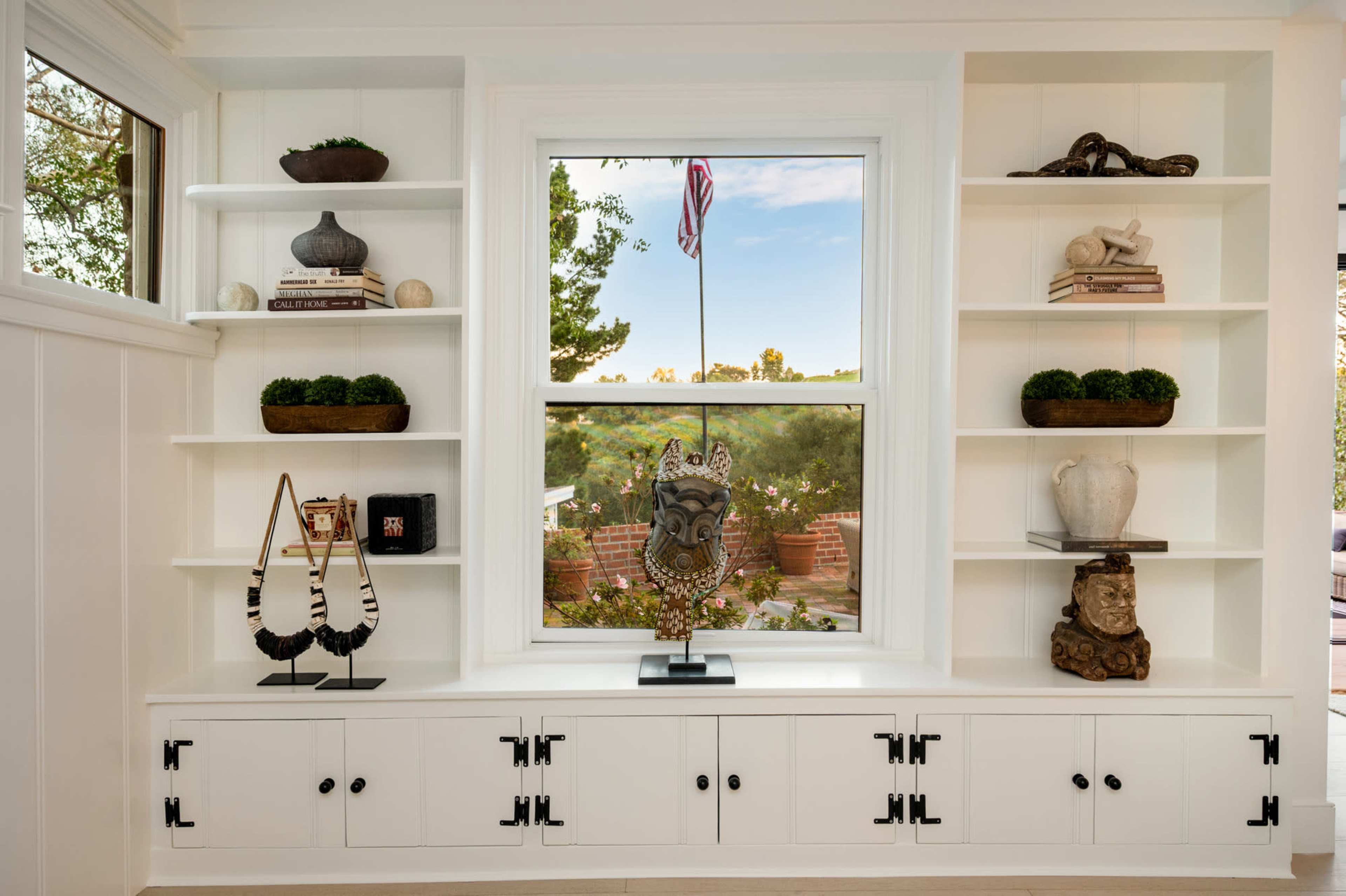 A built-in shelving unit features decorative items and a large window offering a view of greenery and an American flag outside.