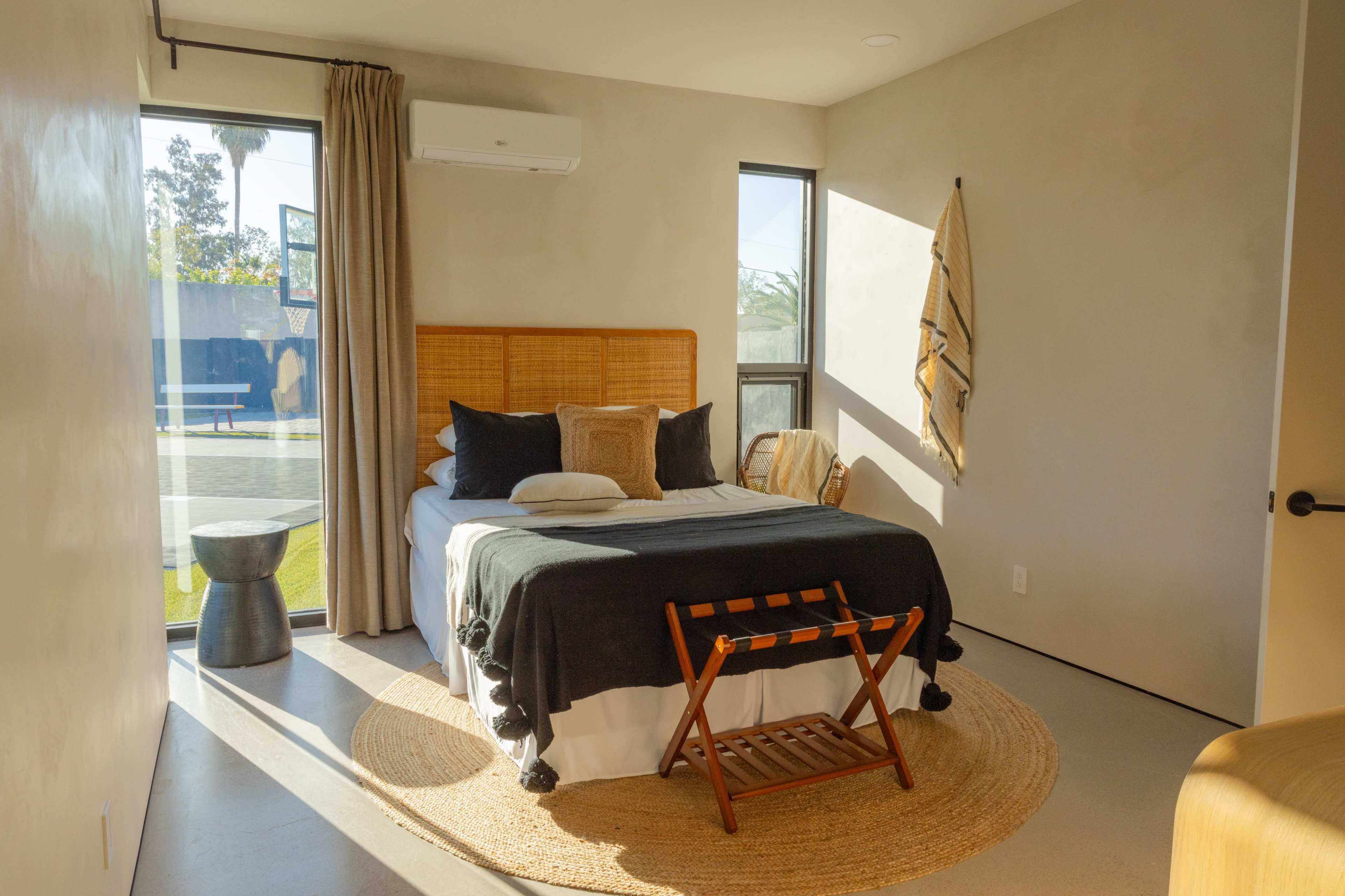 A minimalistic bedroom featuring a bed with dark bedding, a wooden headboard, a round rug, and large windows allowing natural light to enter.
