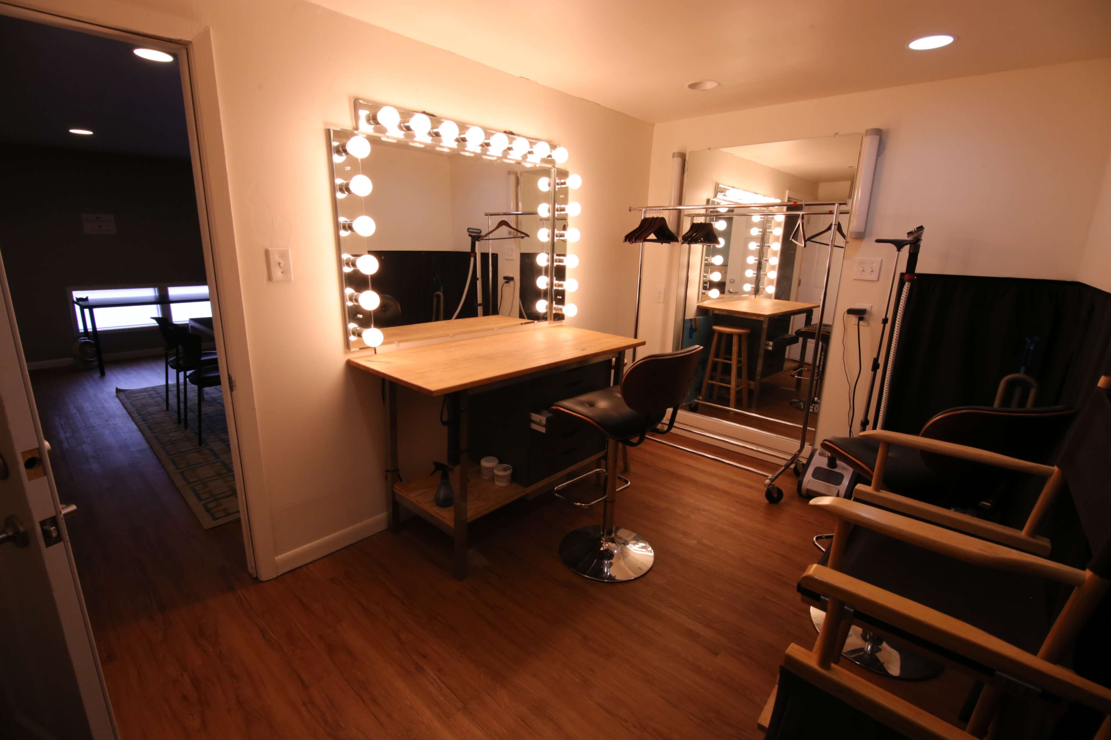 A well-lit dressing room featuring a large mirror surrounded by lights, a wooden table, and a clothing rack.
