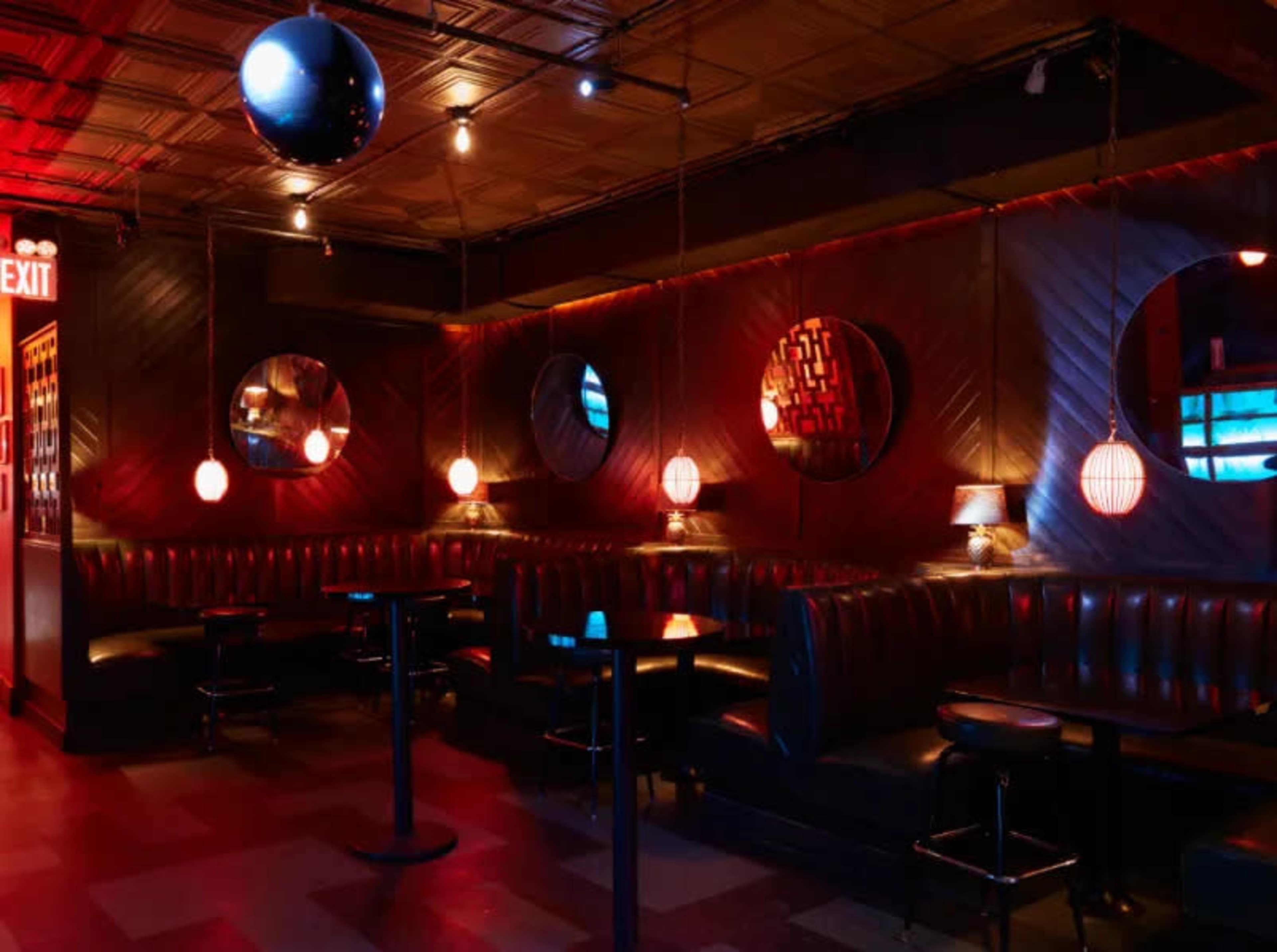The image shows a dimly lit bar with round mirrors and ambient lighting, featuring black leather seating and small tables.