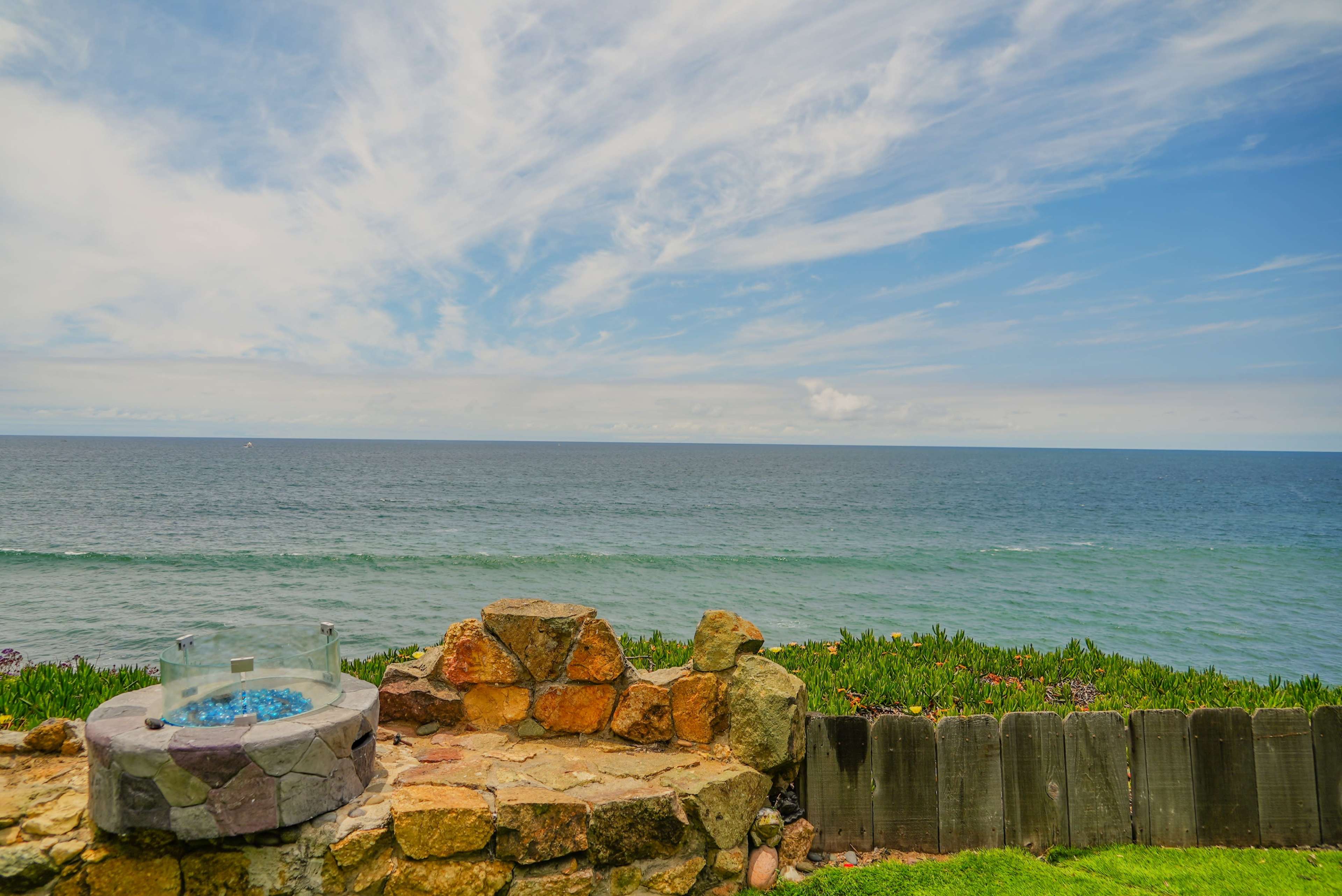 A stone structure with blue glass sits by the edge of a grassy area overlooking a calm ocean under a mostly cloudy sky.