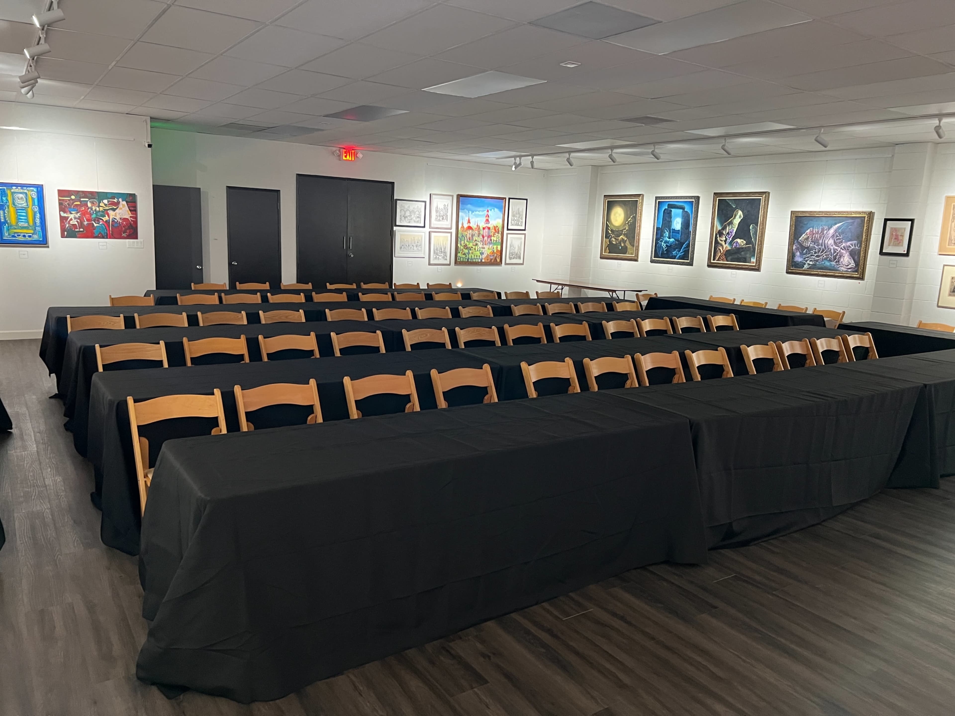 The image shows a large room set up for an event with rows of tables covered in black tablecloths and framed artwork displayed on the walls.