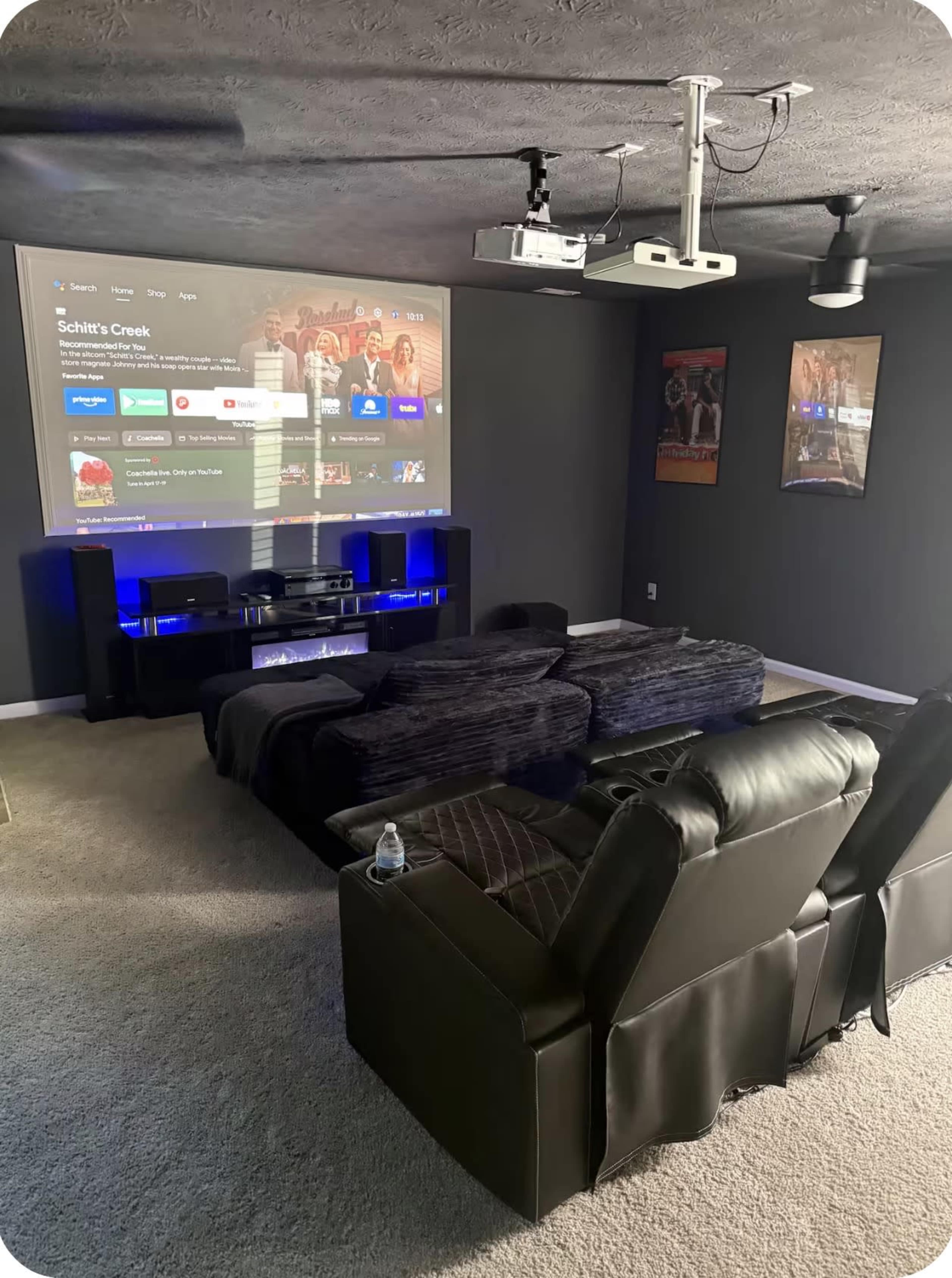 The room features a home theater setup with black leather reclining sofas, a projector displaying a streaming service interface on the wall, and a media console with speakers and ambient lighting.