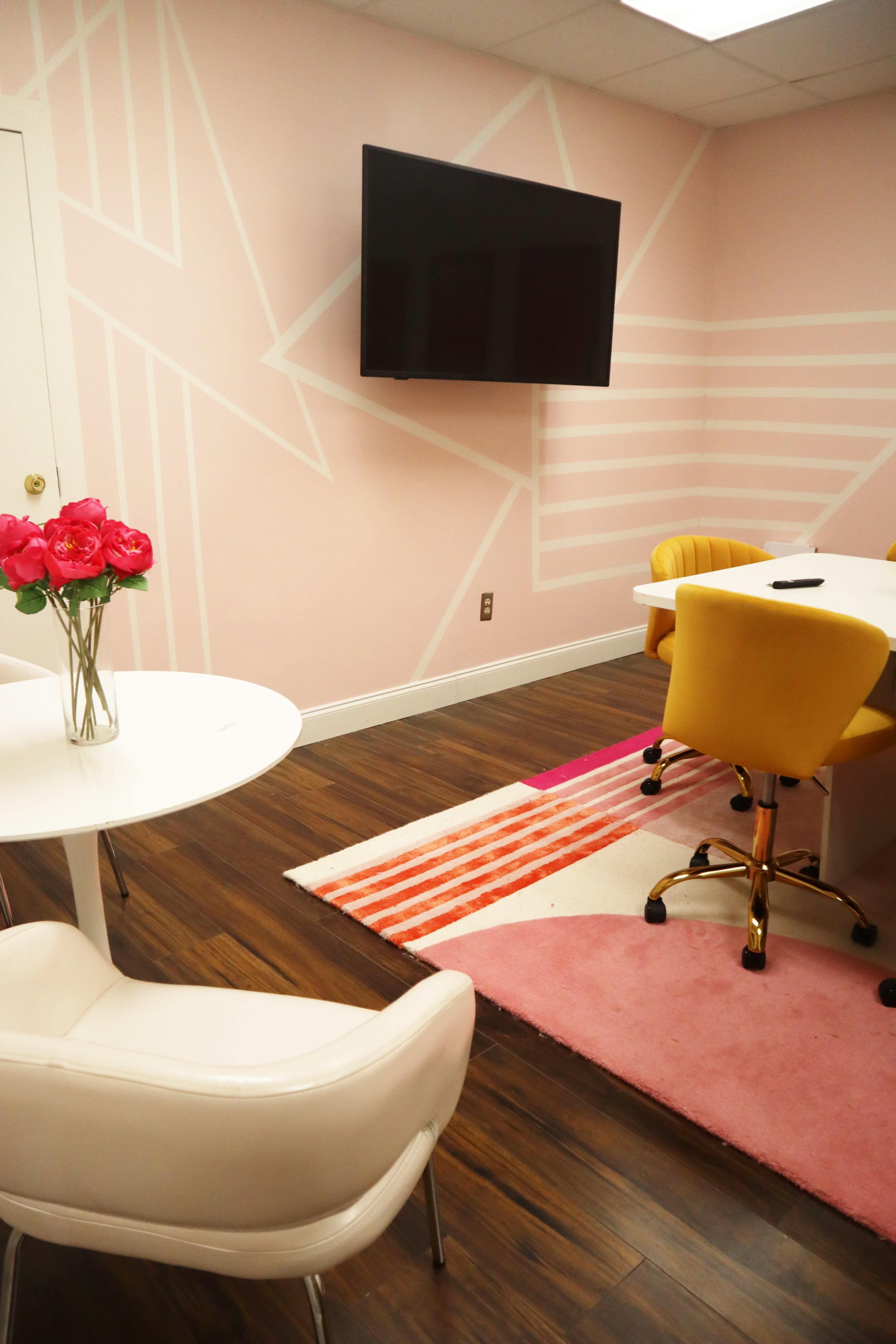 A meeting room with pink walls, a large television, a white table with flowers, and yellow chairs on a multi-colored rug.