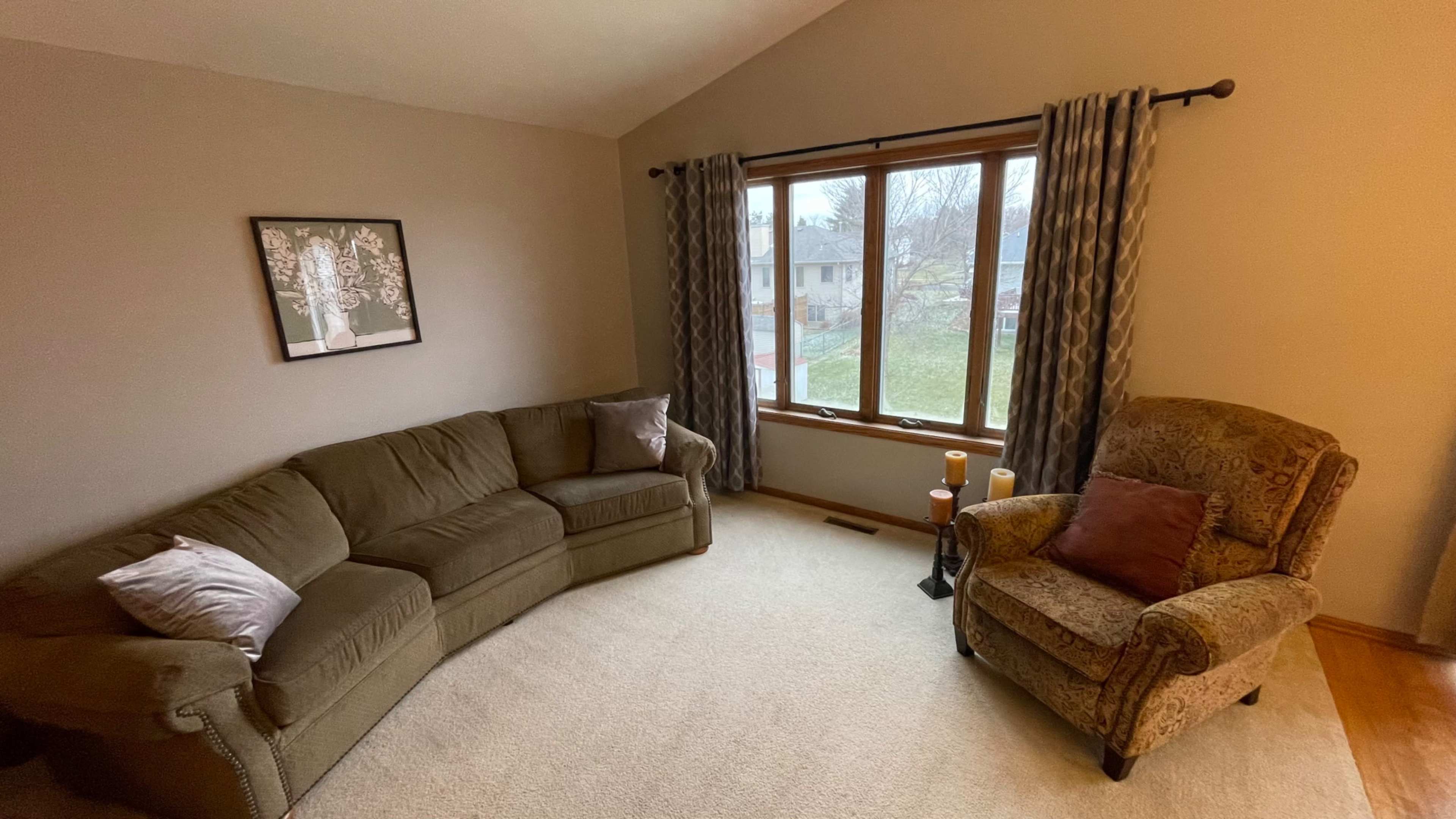 A cozy living room features a curved sofa, an armchair, and large windows with drapes overlooking a yard.