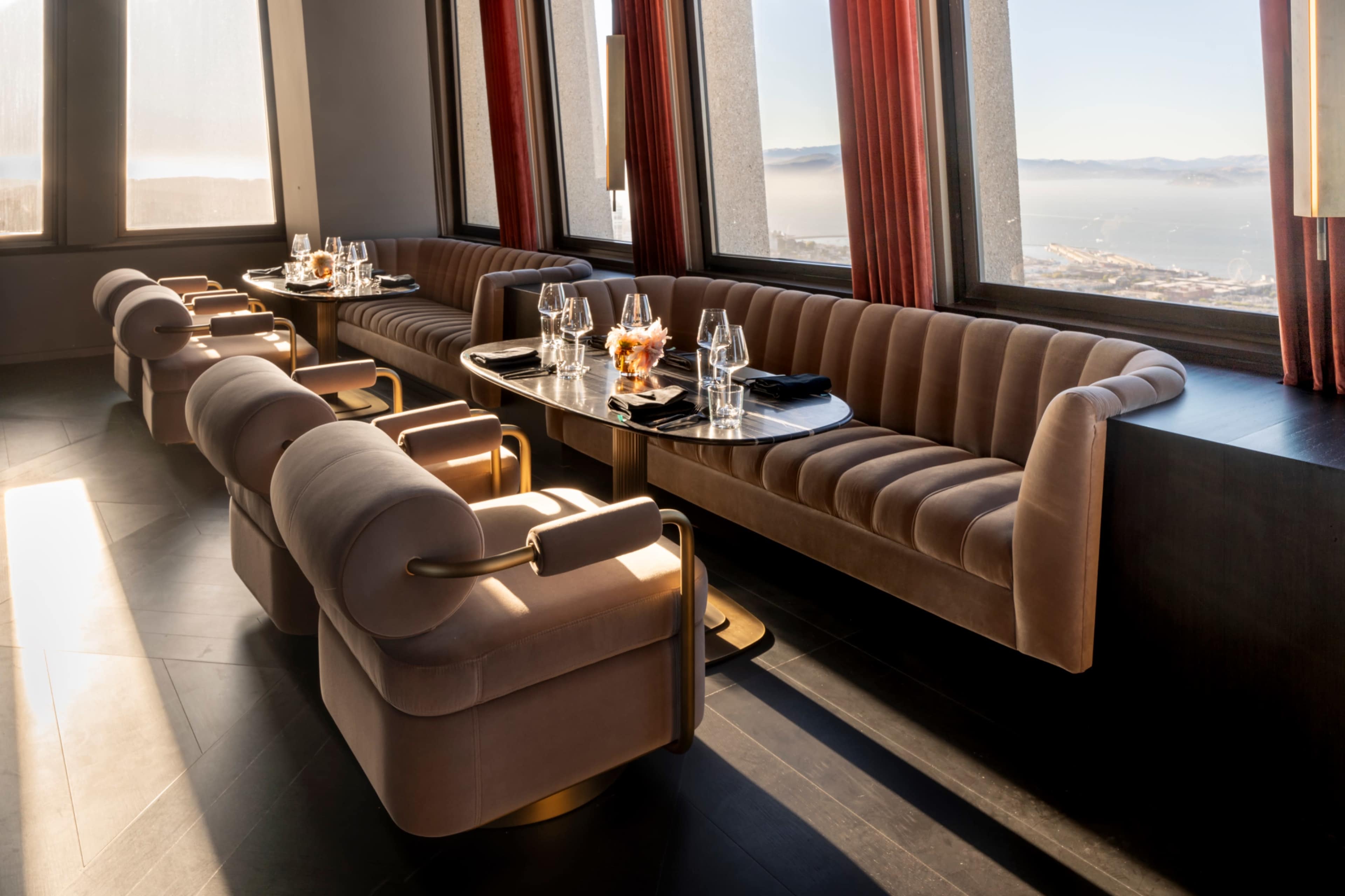 The image shows a modern restaurant interior with curved upholstered seating and elegantly set tables, featuring large windows that provide a view of the landscape.