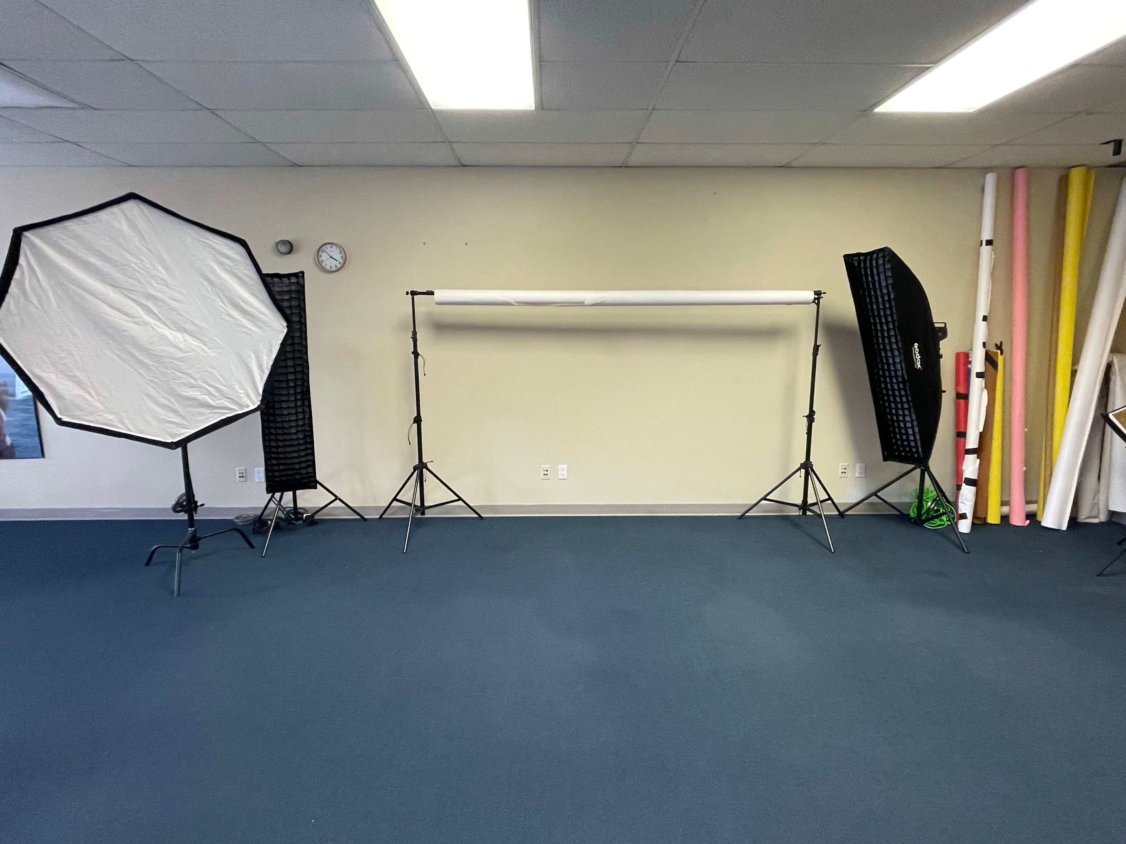 A photography studio with two softbox light setups, a backdrop holder with a white roll of backdrop paper, and various colored foam boards leaning against the wall.