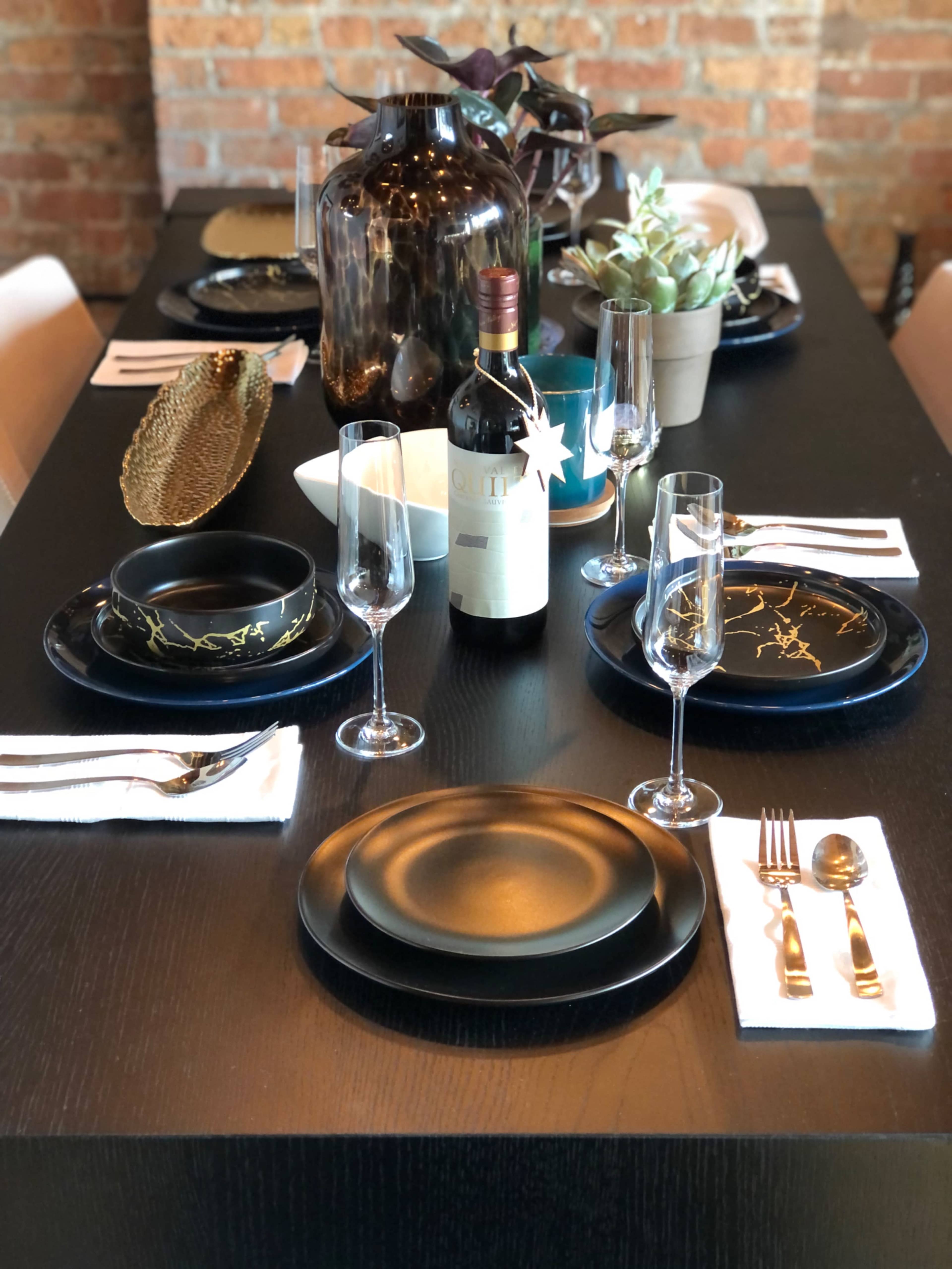 A dining table is set with black plates, wine glasses, and a bottle of wine, surrounded by decorative plants and a striking backdrop of exposed brick.