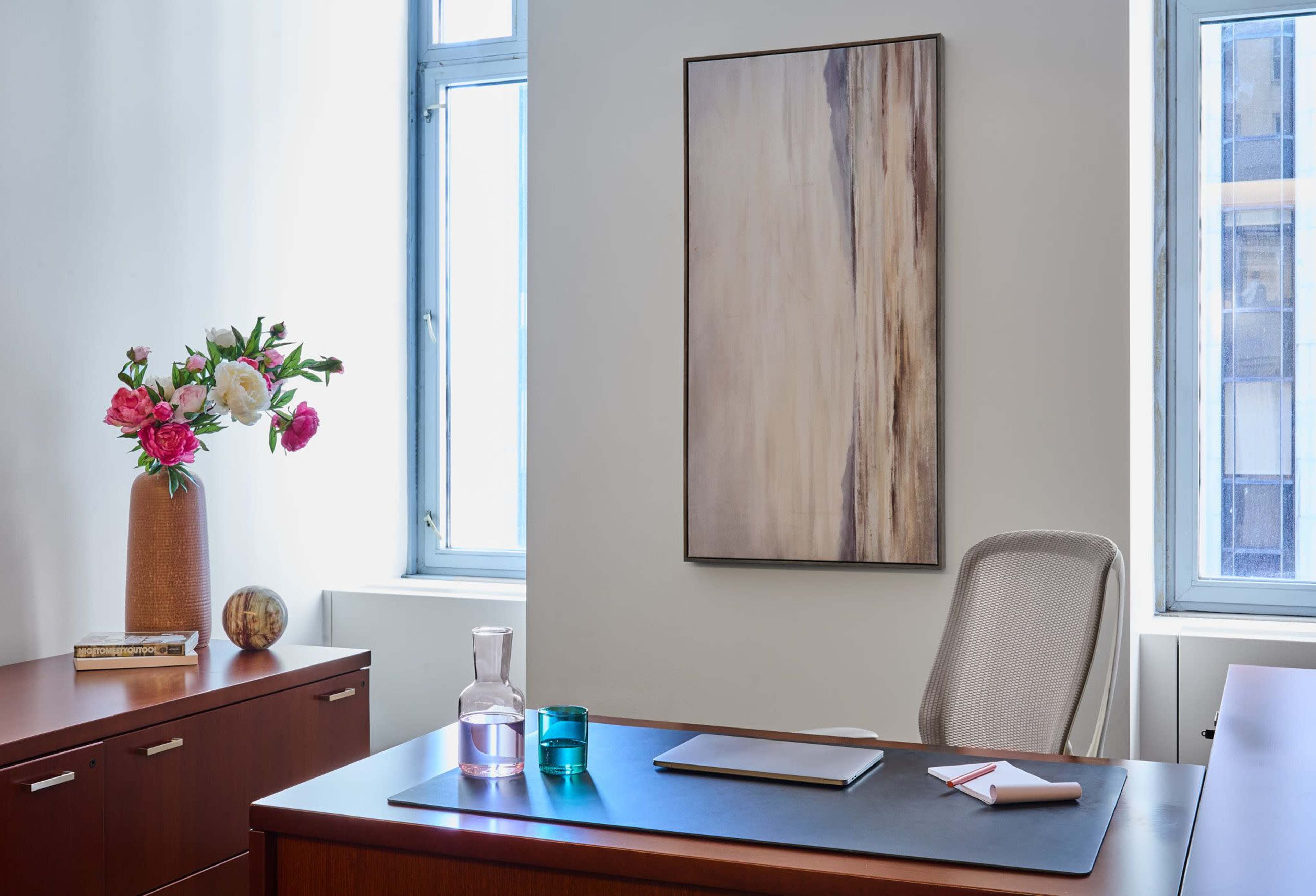 The image shows a modern office space with a wooden desk, a chair, a large piece of wall art, and a vase of flowers on a nearby cabinet.