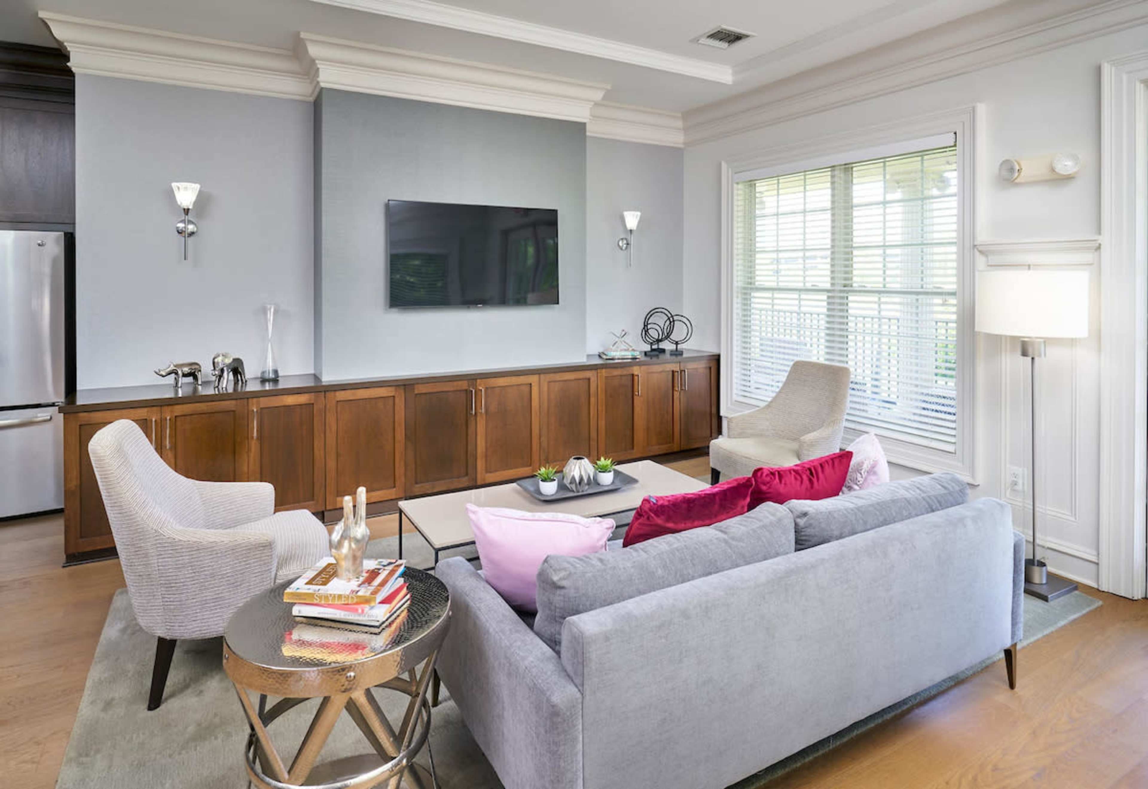 A modern living room features a gray sofa, two armchairs, a round side table, and a wall-mounted television against a backdrop of light-colored walls and a large window.