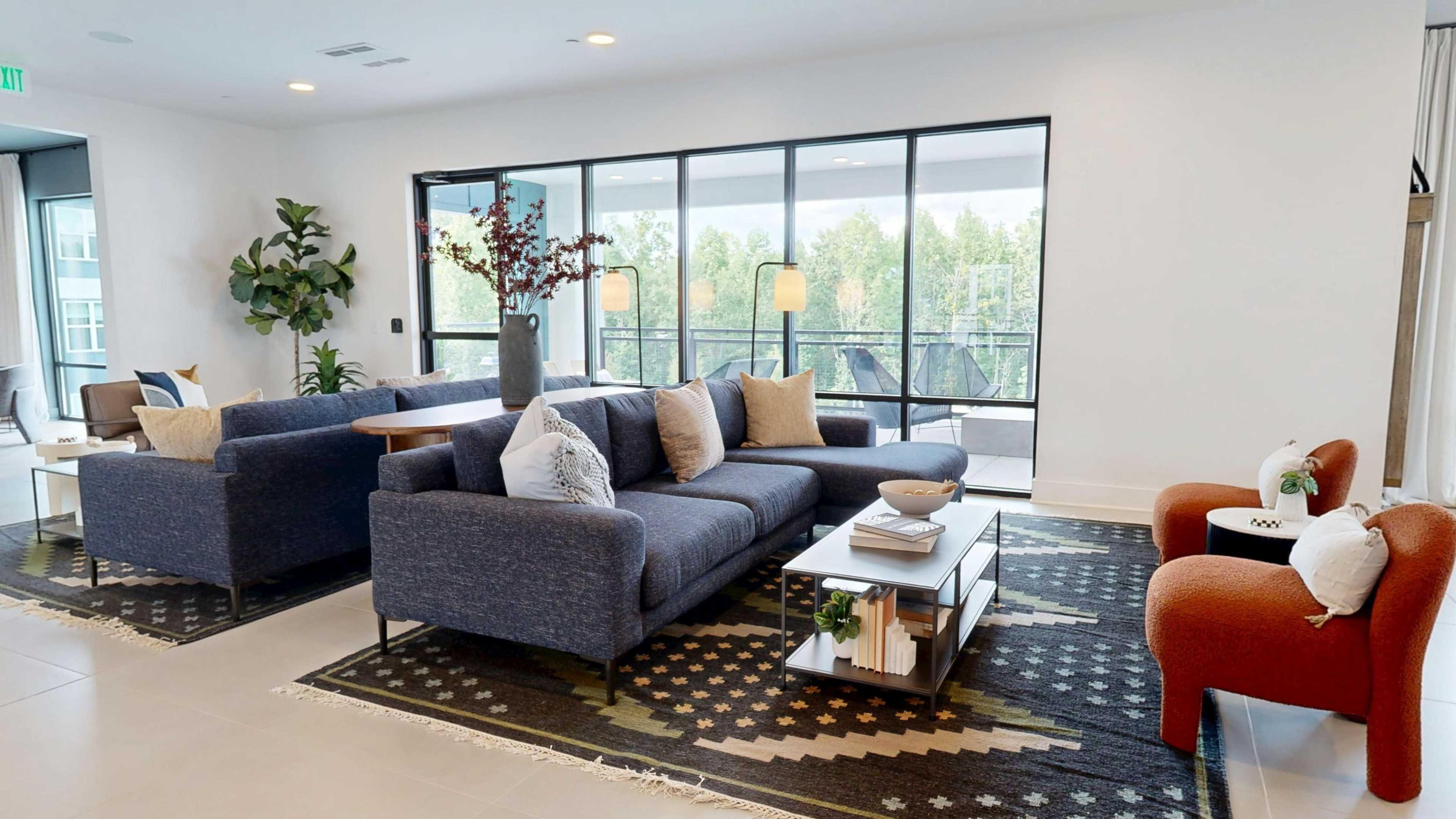 The image shows a modern living room with a blue sectional sofa, a coffee table on a patterned area rug, and large windows overlooking greenery.