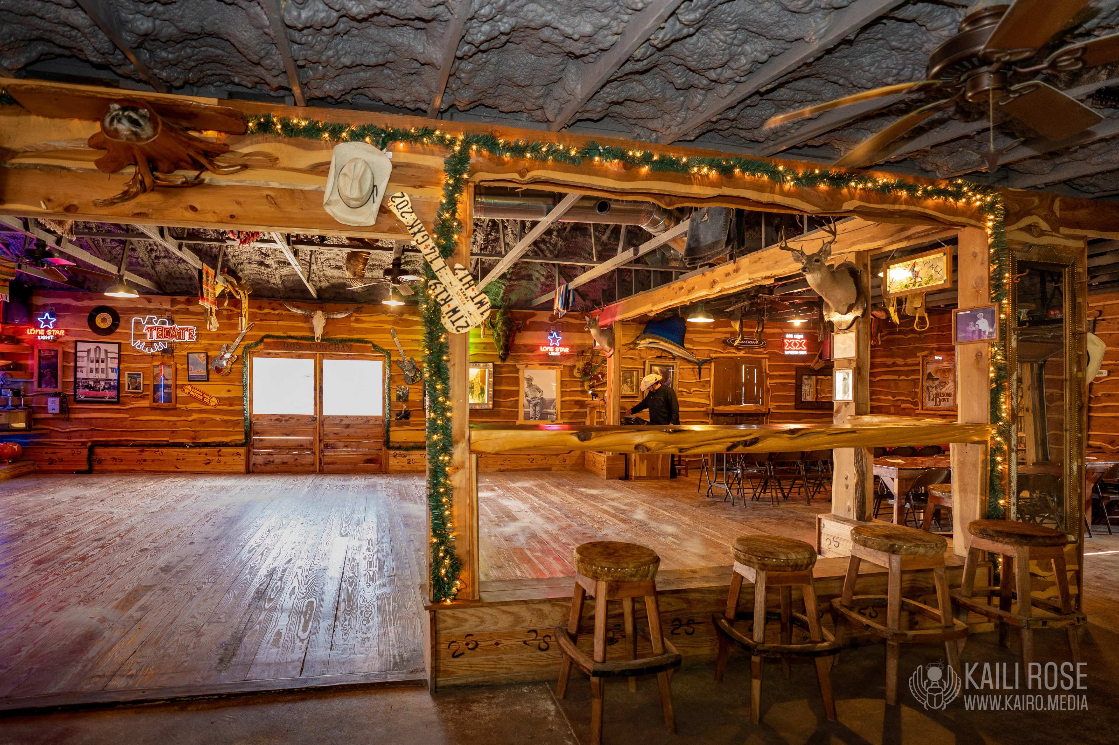 A rustic wooden interior of a bar area with a spacious dance floor, illuminated by colorful neon lights and decorated with various memorabilia.