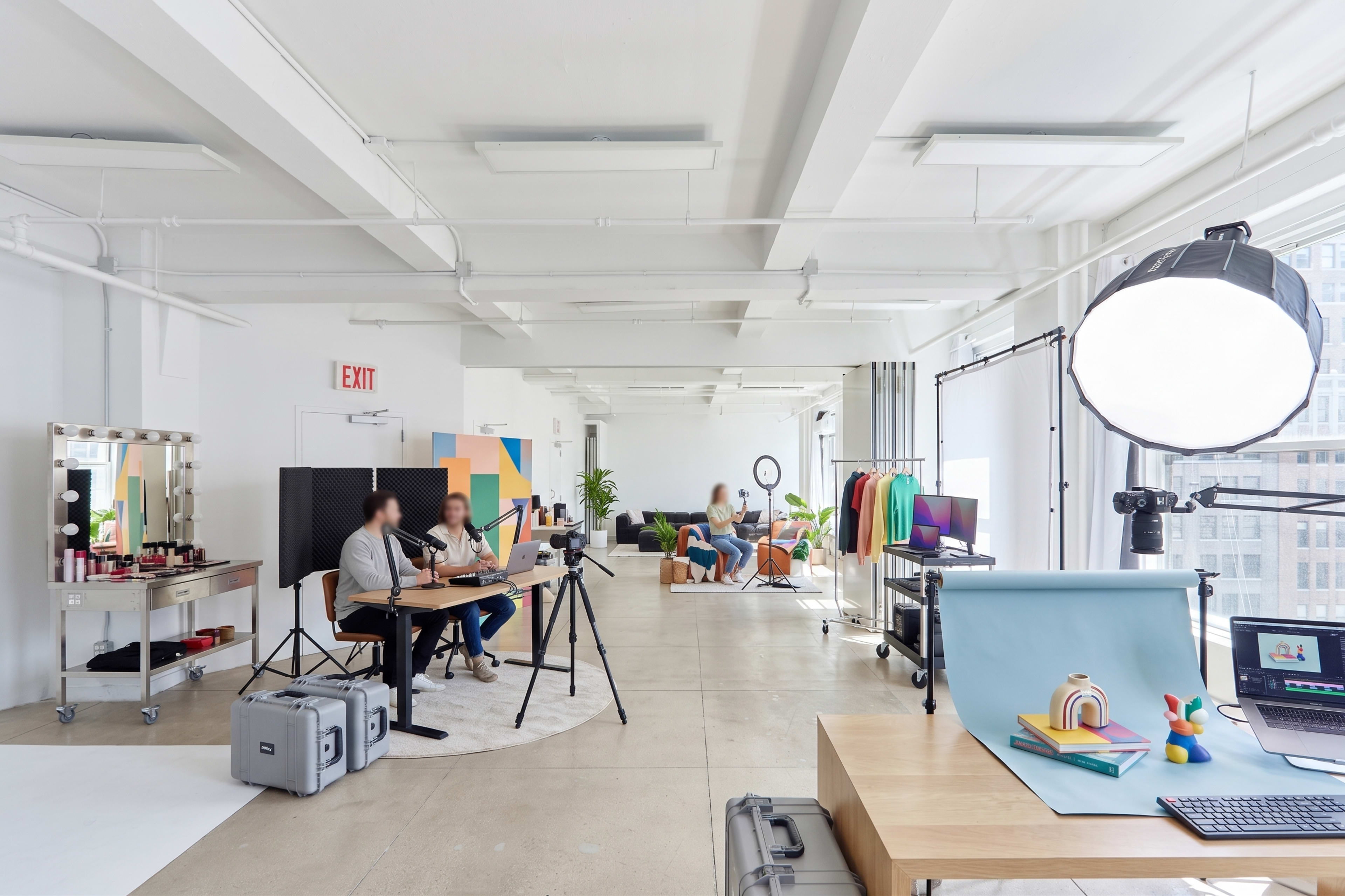 The image depicts a brightly lit studio space featuring a table with equipment, two individuals seated at the table, and various creative items spread throughout the room.