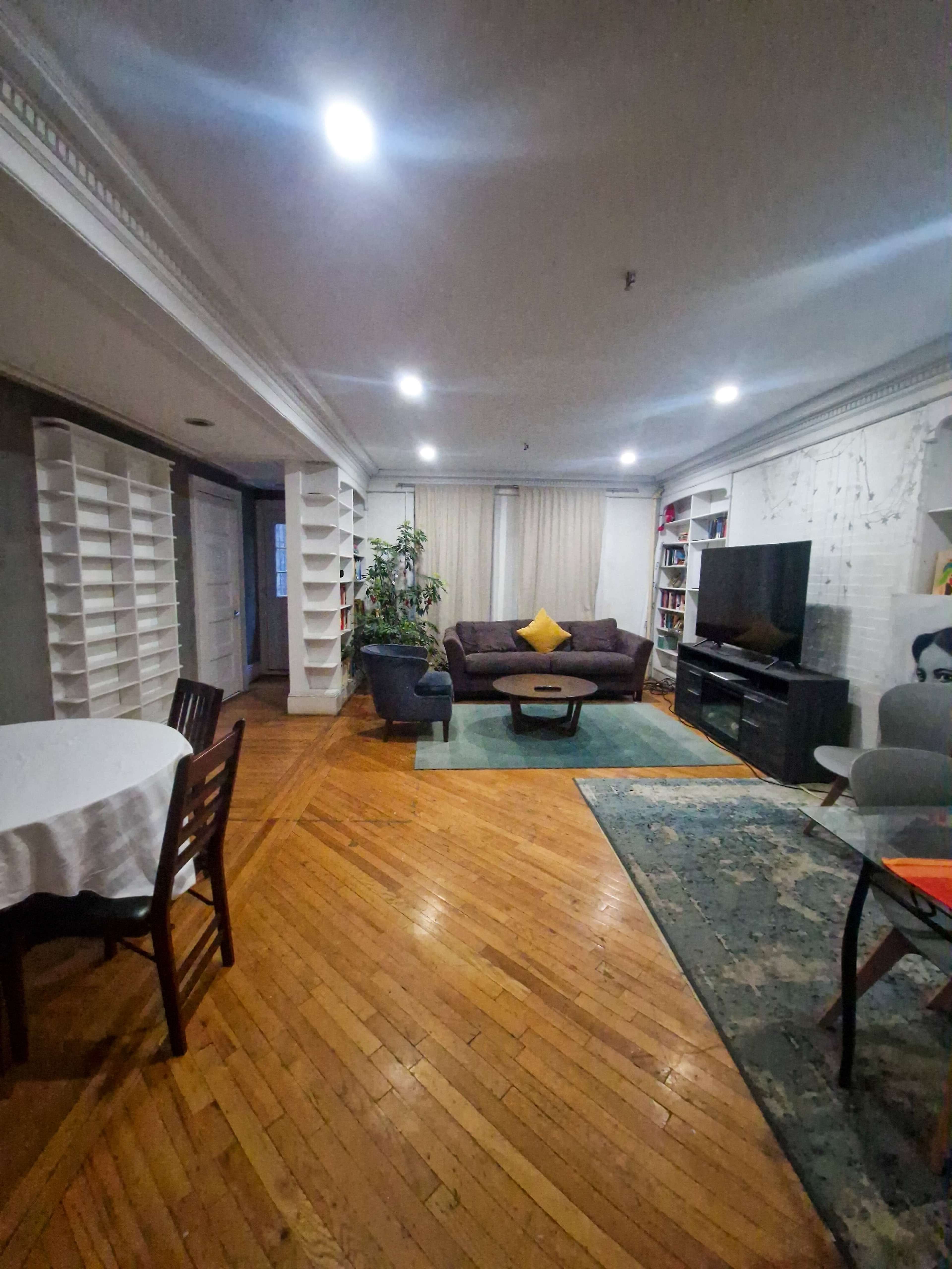 The image shows a spacious living area with hardwood floors, featuring a seating arrangement, a television, bookshelves, and a dining table.