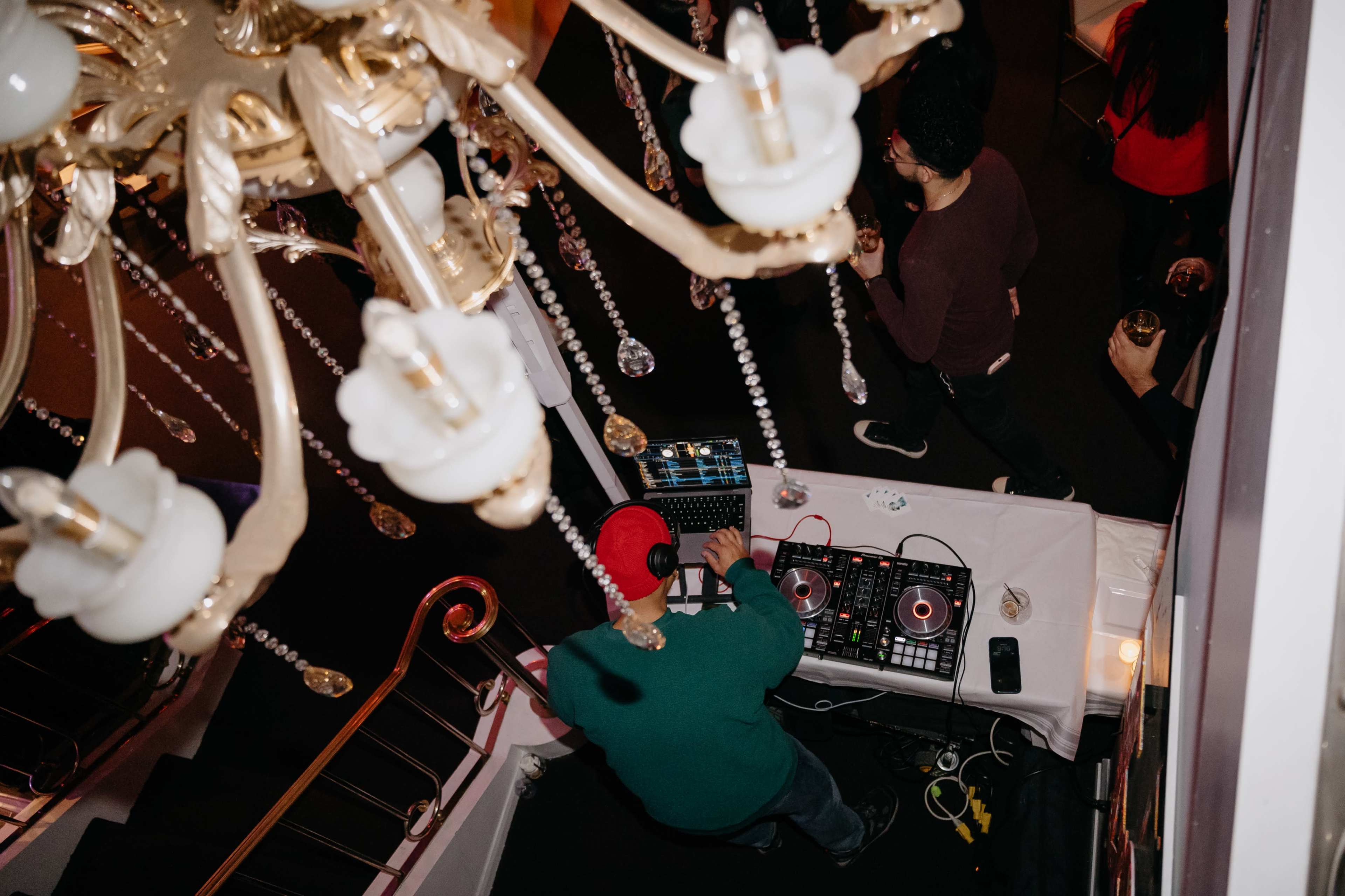 A DJ wearing a red hat mixes music at a table on a staircase while guests socialize in the background.