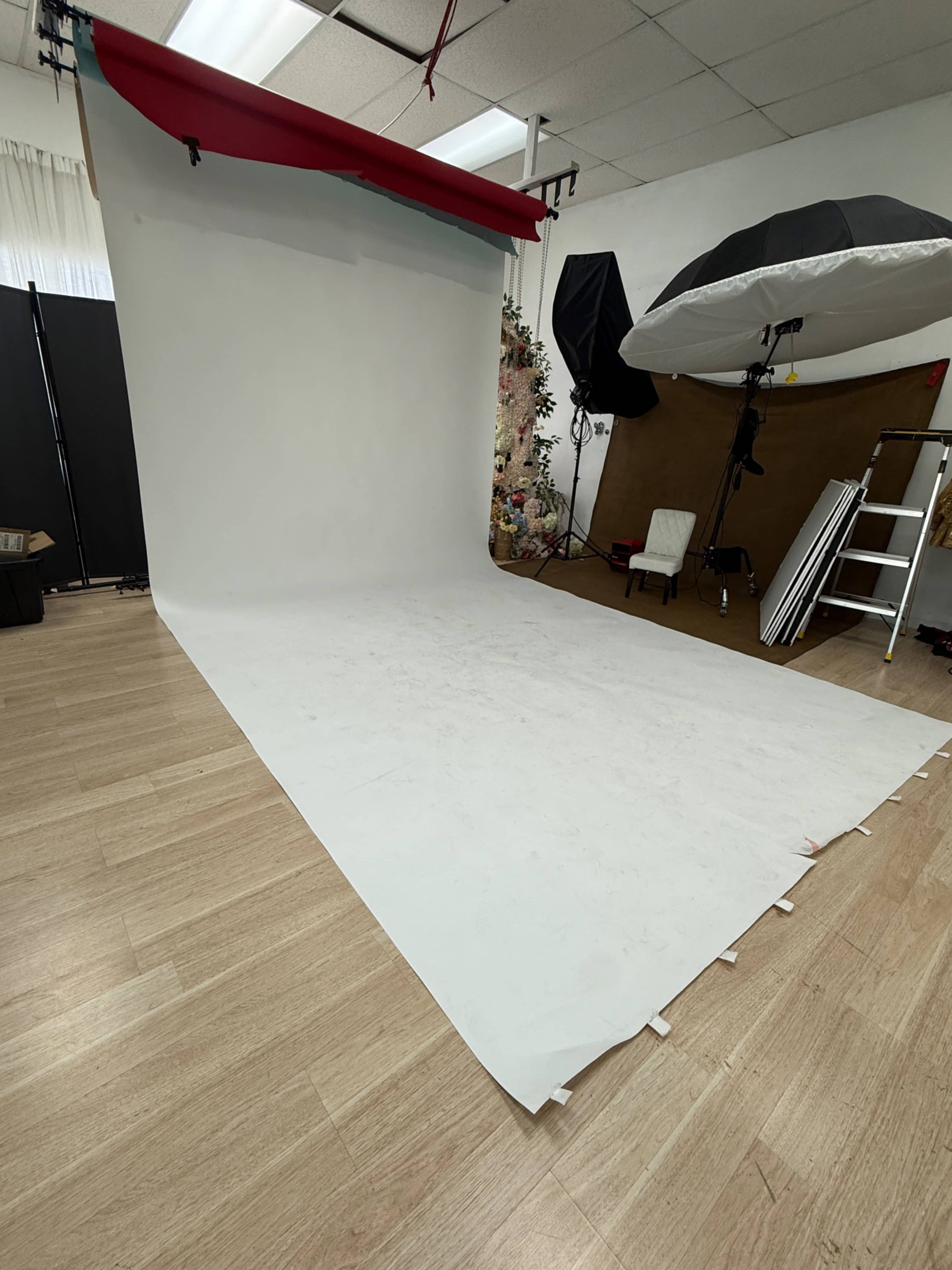 The image shows a photography studio with a large white backdrop, softbox lights, a ladder, and various props arranged around the space.