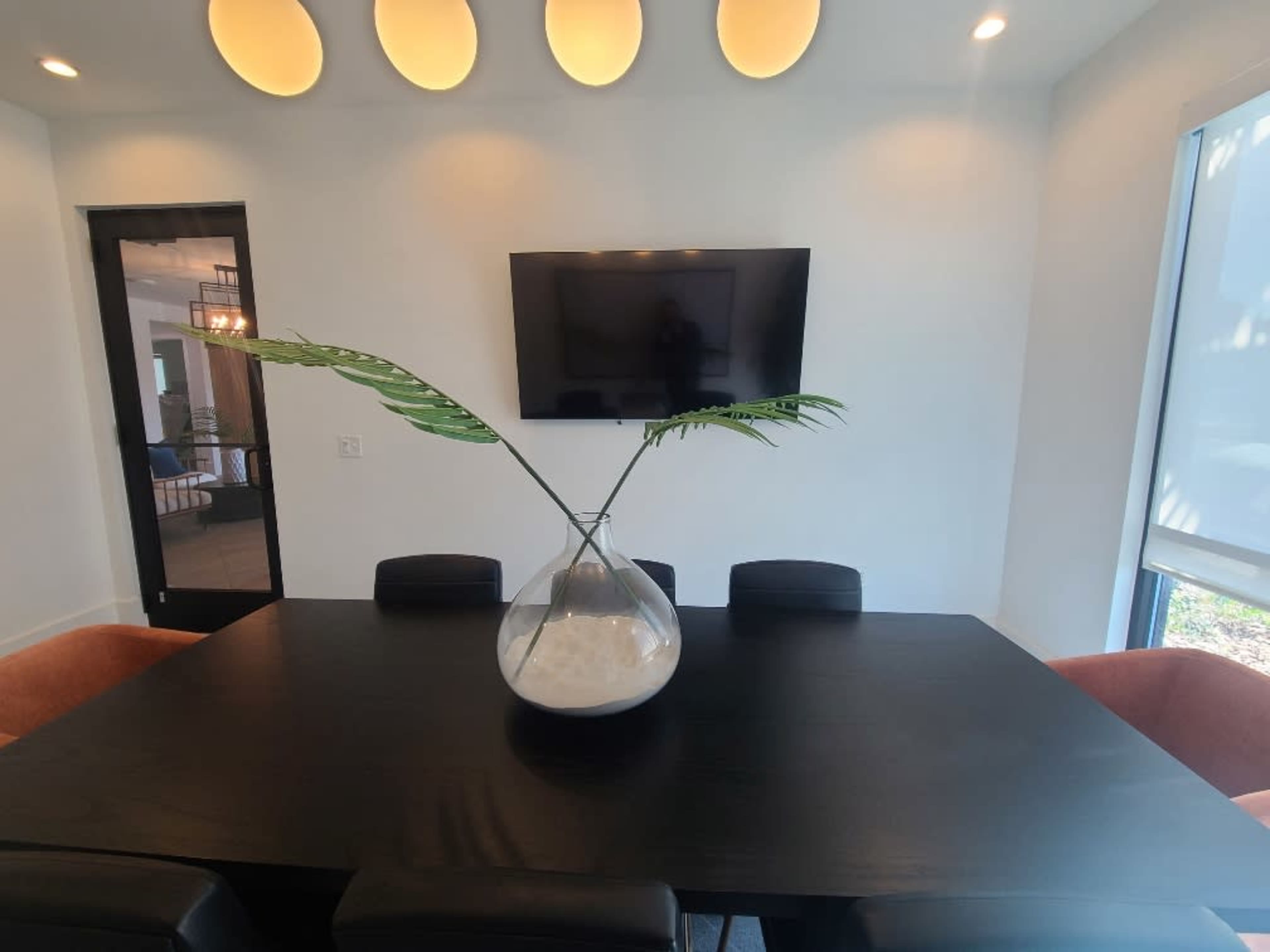A modern dining room features a black table with a large glass vase containing a decorative plant, complemented by a wall-mounted television and illuminated ceiling lights.