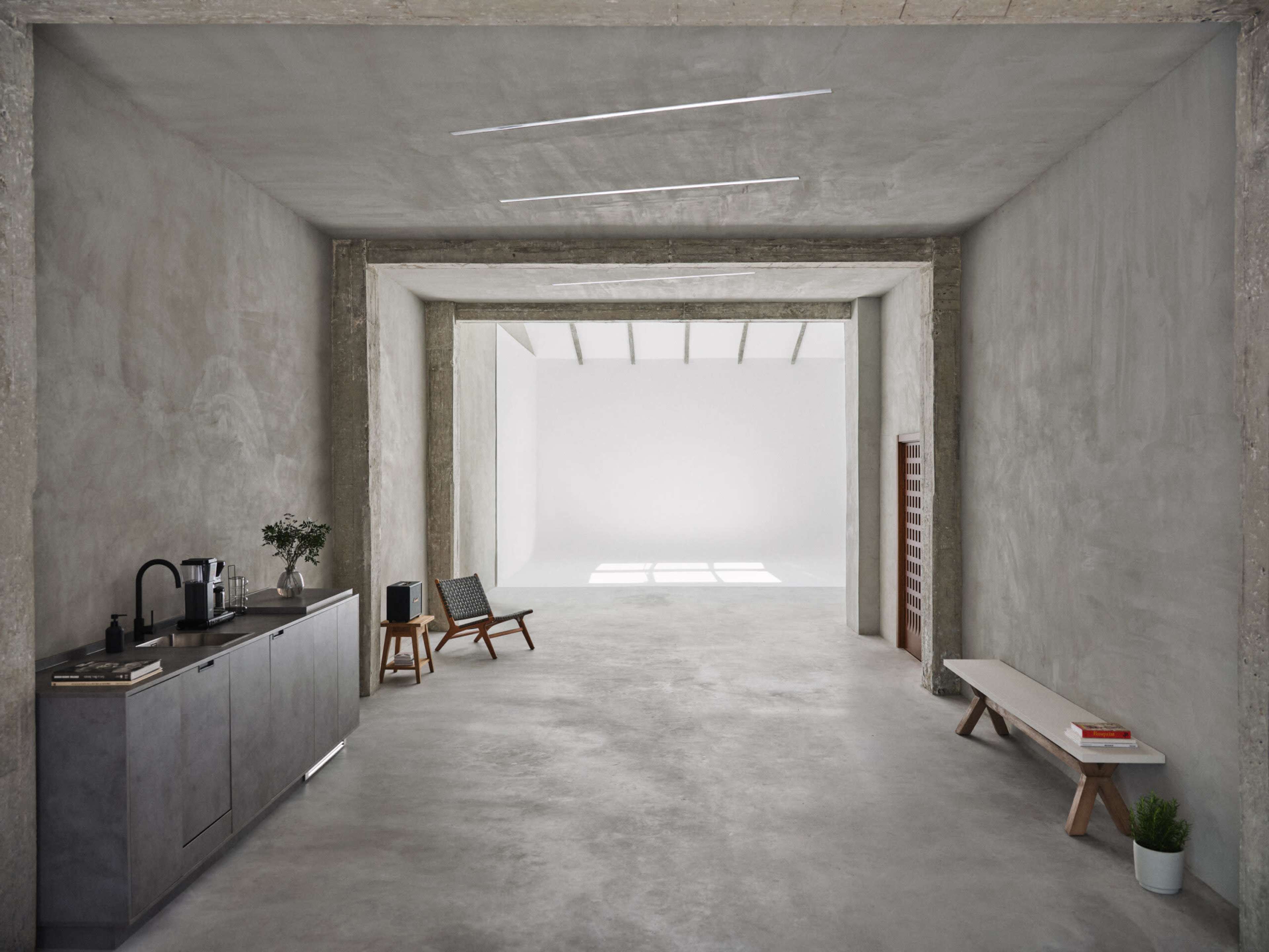 The image shows a spacious, minimalistic interior with concrete walls, an open layout, and a modern kitchenette on one side.