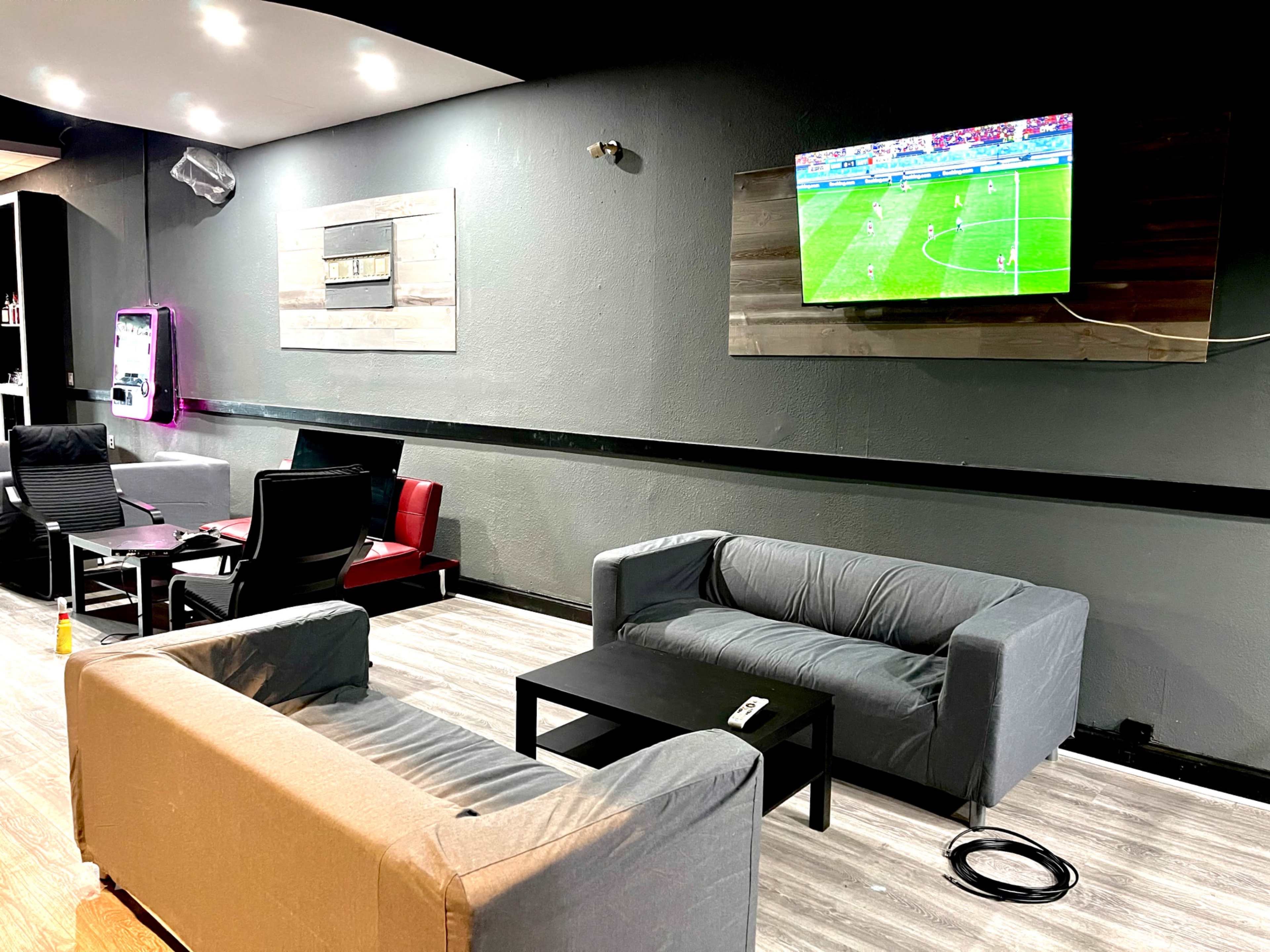 The image shows a lounge area with gray and red seating, a coffee table, and a television displaying a soccer match on the wall.