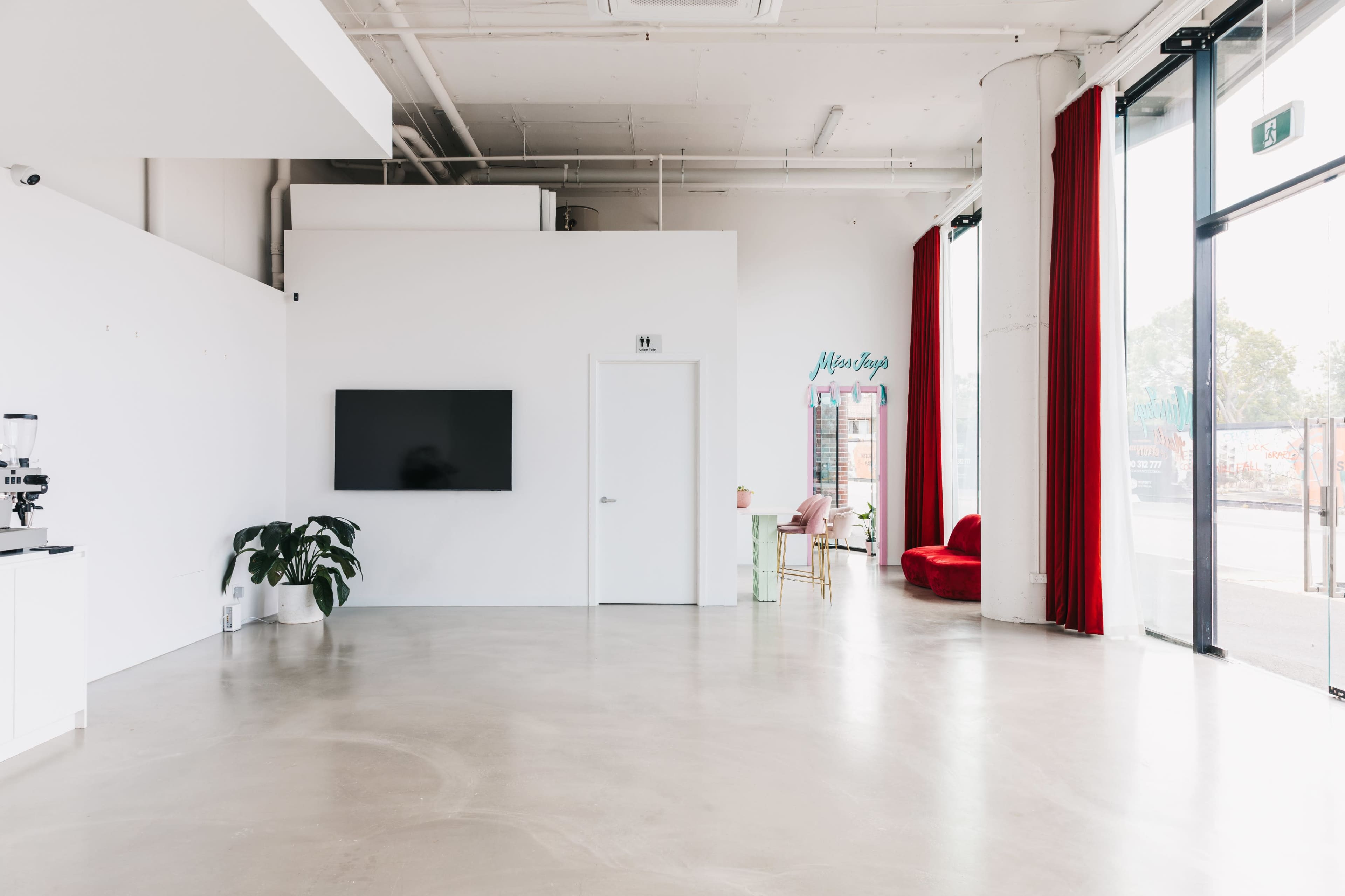 The image shows a spacious, minimalist interior with white walls, large windows adorned with red curtains, a green plant, and a seating area with a red chair.
