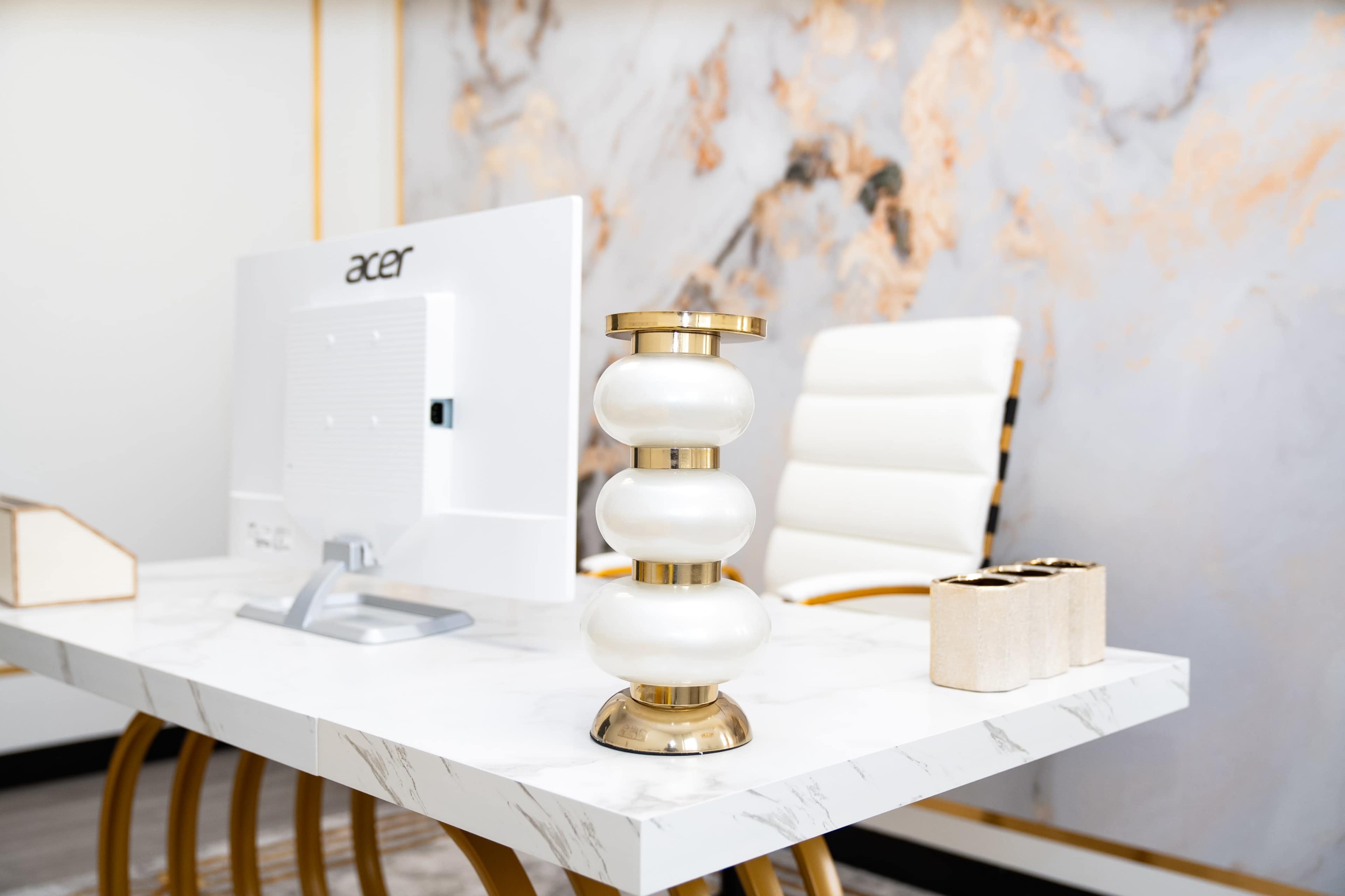 A modern office desk features a sleek computer monitor, a decorative lamp with a gold base, and a holder for pens against a marbled wall.